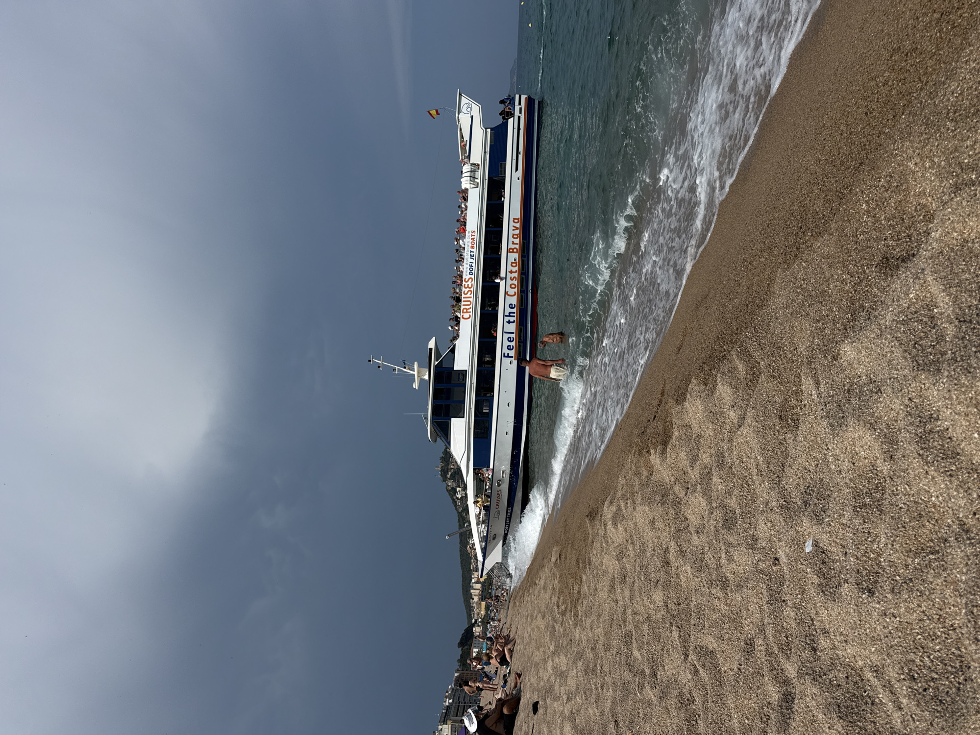 This media file captures a bright, sunny day at a beach in Blanes, Spain. The scene is dominated by a large, multi-deck passenger boat that appears to be either docking or departing near the shore. The boat is white with blue trim and features the text "CRUISES DOFI JET BOATS" and "Feel the Costa Brava" printed prominently on its side. Numerous passengers are visible on the decks of the boat, enjoying the excursion.
In the foreground, the sandy beach is visible with gentle waves lapping at the shore. Several people are present on the beach, some sunbathing, others wading into the water. The water is a clear blue, and the sky is a bright, overcast grey, suggesting it might be later in the day or a partially cloudy afternoon. The overall atmosphere seems relaxed and summery, indicative of a tourist destination. In the background, a coastal town with buildings nestled on a hillside can be seen, confirming the location as a Mediterranean seaside resort.
