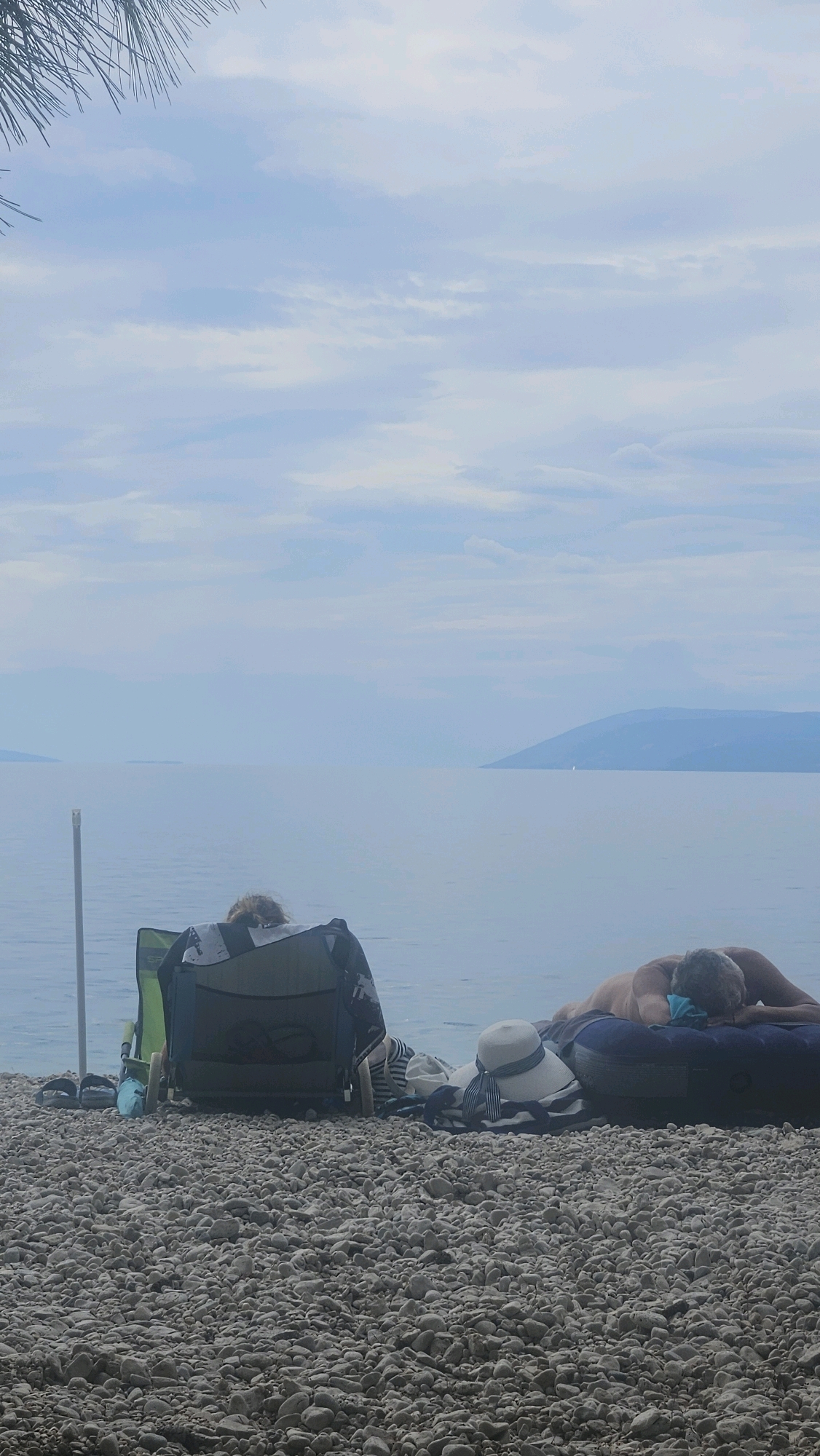 This is a vertical, eye-level, full shot of a pebbled beach in Općina Podgora, Croatia. The scene is dominated by a calm sea and a hazy, overcast sky. In the foreground, a person sits in a green camping chair facing away from the camera, with a dark patterned towel draped over the chair. To the right of the chair is a large, blue inflatable raft with a person lying face down on it, with their head resting on a blue pillow. Next to the inflatable raft is a white sun hat with a blue and white striped ribbon. In the distance, the sea stretches to a hazy coastline with mountains. The overall atmosphere is tranquil and relaxed.