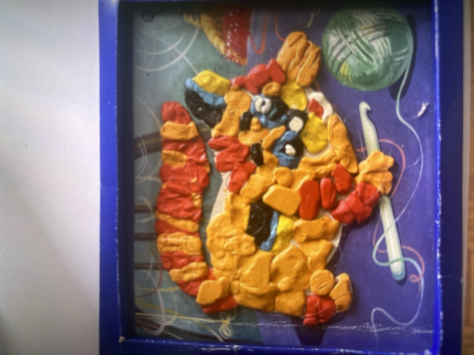The image shows a colorful, textured artwork composed of what appears to be clay or play-doh. The artwork is framed by a dark blue border and is set against a purple background with abstract line drawings. The central subject of the artwork is a creature with a segmented, red and yellow body, resembling a caterpillar or a dragon. It has multiple limbs and a detailed head with black and blue elements that could represent eyes or facial features. To the lower left of the creature is a light blue crochet hook with a white handle, positioned as if it's part of the scene's narrative or simply an artistic element. To the lower right is a ball of green yarn with visible texture. The background features swirling lines in white, pink, and yellow, suggesting movement or a decorative pattern. The overall impression is whimsical and handmade.
The image does not provide any visual cues or context that would allow for the identification of its location in the city of Kerava, Finland.