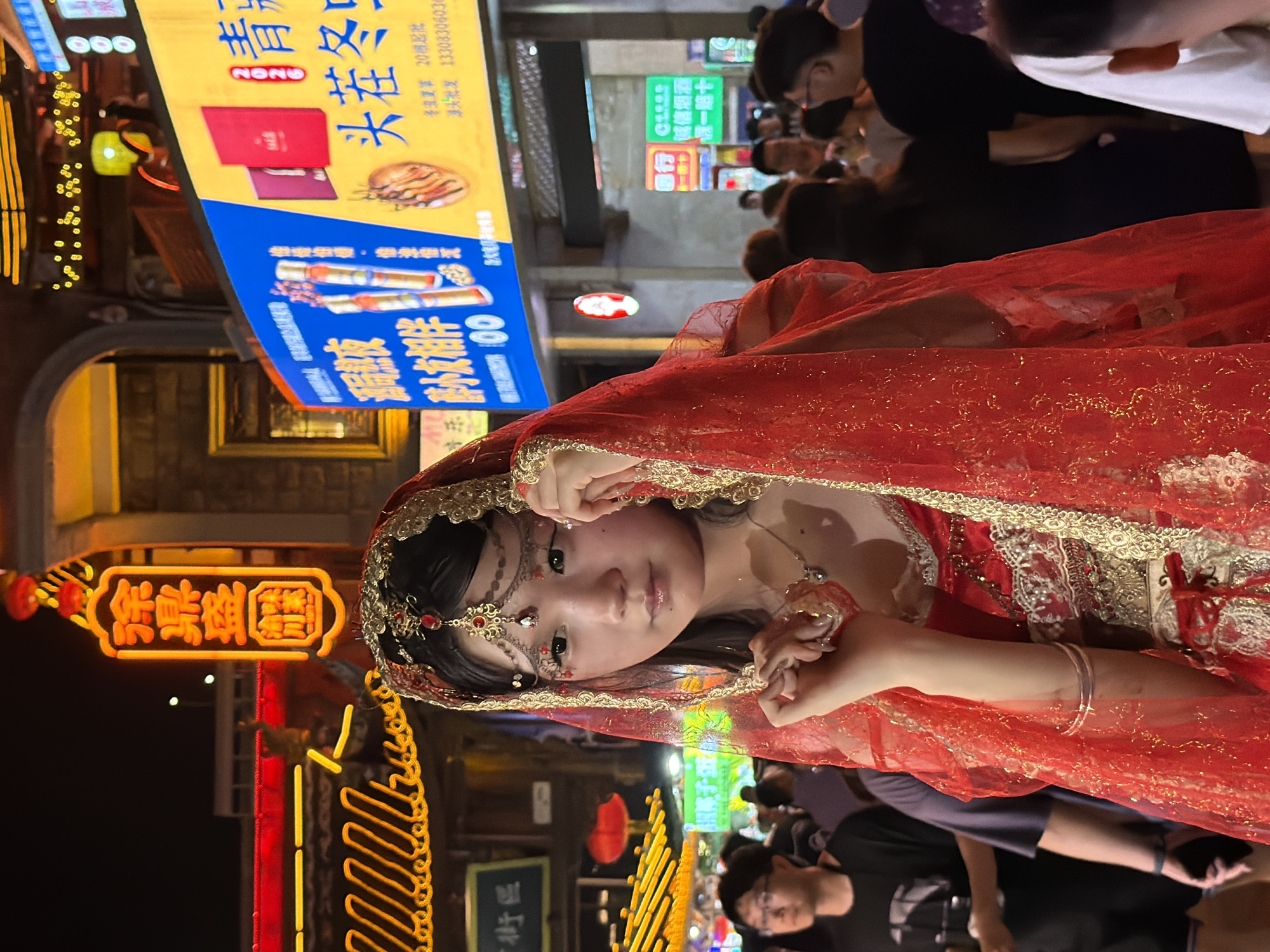 A young woman in a red and gold ornate garment with a veil is the central focus of the image, positioned in the foreground against a blurred backdrop of a bustling night market. She has dark hair and is adorned with jewelry on her forehead and around her neck. Her hands are near her face, and she appears to be looking slightly away from the camera. The background depicts a lively street scene with neon signs, traditional Chinese architecture, and a crowd of people. The overall lighting suggests it is nighttime, with artificial lights illuminating the market stalls and advertisements. The woman's attire suggests a cultural or ceremonial context, possibly a performance or festival. The location is identified as Yuzhong District, China, implying an urban setting characteristic of such areas.
