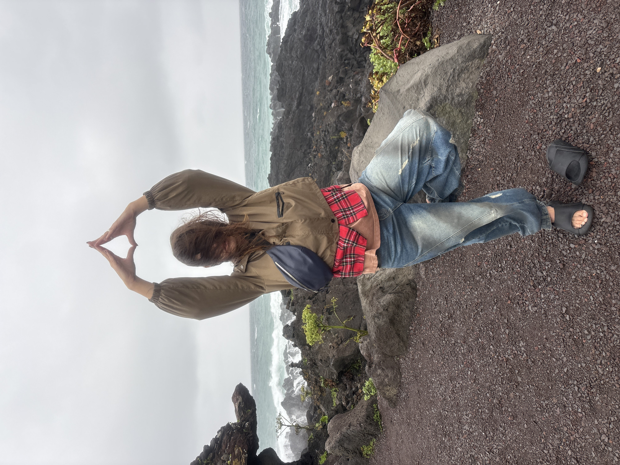 In Seogwipo-si, South Korea, a person is captured in a low-angle, vertical shot, sitting on a large rock next to a dark, gravel path. The person, wearing a khaki jacket, a red and black plaid shirt tied around their waist, and distressed blue jeans, is making a triangular shape with their hands above their head. A navy fanny pack is worn across their chest. Black slides are on their feet, with one detached on the ground.
The setting is a rocky coastline with crashing waves against dark volcanic rocks. The ocean is a turbulent green-blue under an overcast sky, suggesting a cloudy or misty day. Small green plants are growing among the rocks. The overall mood is somewhat dramatic, emphasized by the rugged landscape and the powerful ocean. The time of day appears to be daytime due to the ambient light, but the overcast conditions prevent distinct shadows. No visible text is present in the image.