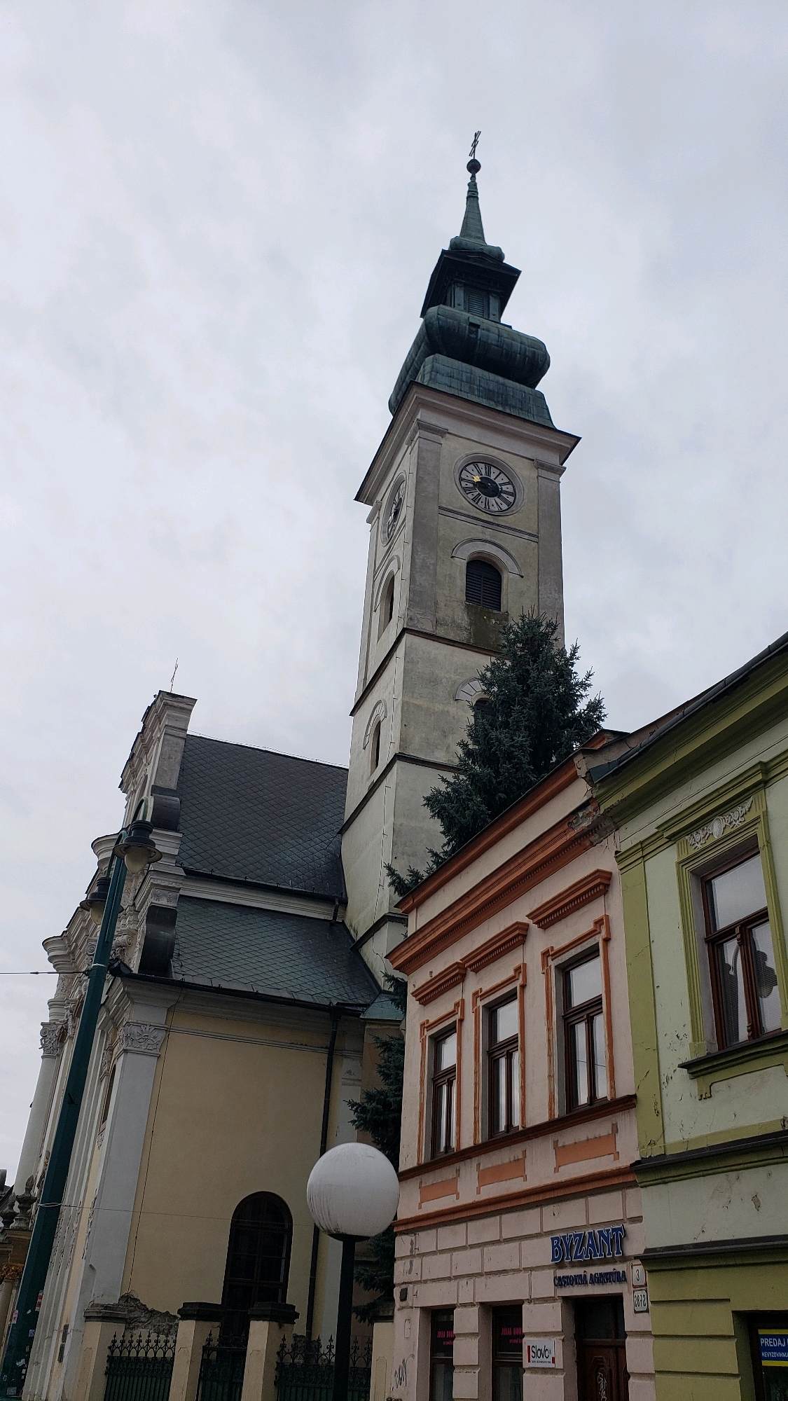 This is a vertical, medium-range, eye-level shot of a clock tower and adjacent buildings in Prešov, Slovakia. The clock tower is on the left, and it is a pale grey structure with a dark green, onion-shaped spire. The tower has a clock face with Roman numerals and dark hands. To the right of the clock tower are two buildings. The first building is light orange with decorative trim, and the second building is light green and appears to be an older structure with peeling paint. Both of these buildings have multiple windows with dark frames. In the foreground on the right is a sign that reads "BYZANT" in blue letters above "ČASOVNICTVÁ AGENTÚRA". There is also a sign that reads "PREDAJ NEHNUTEĽNOSTÍ" which means "Real Estate Sales". In the background on the left is a large, pale yellow building with a dark grey, slate-tiled roof and architectural details. A large, white spherical lamp post is visible in the foreground, to the left of the orange building and in front of the yellow building. Several evergreen trees are growing between the clock tower and the orange building. The sky is overcast and a pale grey. The scene is an outdoor urban setting on a cloudy day.