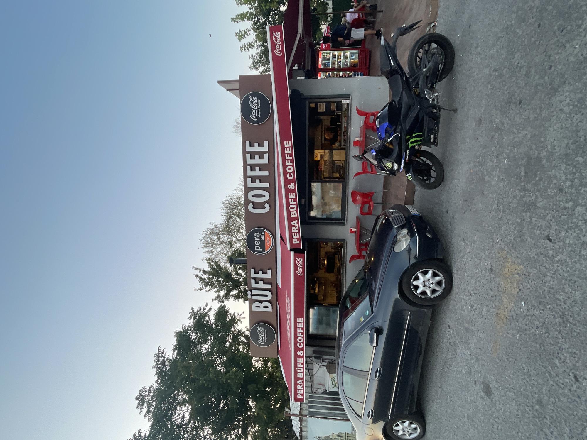 The image depicts a street-level view of a cafe named "PERA BÜFE & COFFEE" in Kadıköy, Turkey. The cafe has a prominent sign displaying "BÜFE" and "COFFEE" in large letters, with Coca-Cola branding visible on awnings and signs. In front of the cafe, a dark-colored sedan is parked to the left, and a black motorcycle is parked to the right. The cafe features outdoor seating with red chairs and tables. A man wearing a backpack is visible standing near a red refrigerator unit at the right side of the cafe. The environment appears to be an urban street with trees in the background and a clear sky.