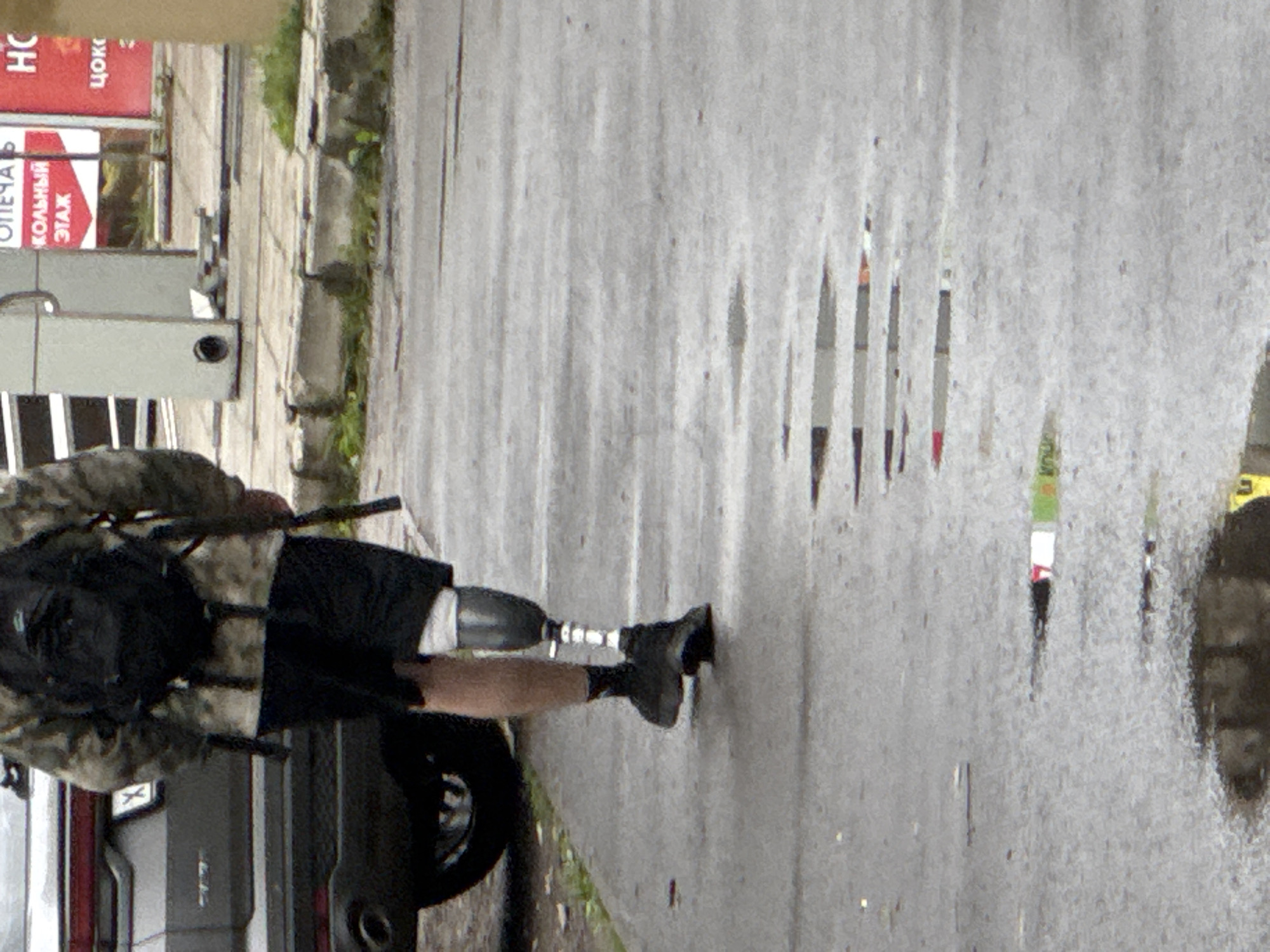 A person with a prosthetic leg walks on a wet asphalt surface in an urban environment. The person is wearing camouflage-patterned clothing, black shorts, and black shoes. A black backpack is worn on their back. To the left of the person is the rear of a dark-colored SUV. In the background, a sidewalk is visible with a light-colored pole and posters. The asphalt surface shows reflections and the presence of puddles, suggesting recent rainfall. Several elongated, thin objects, possibly discarded pens or markers, are scattered on the wet asphalt in a diagonal line behind the person.