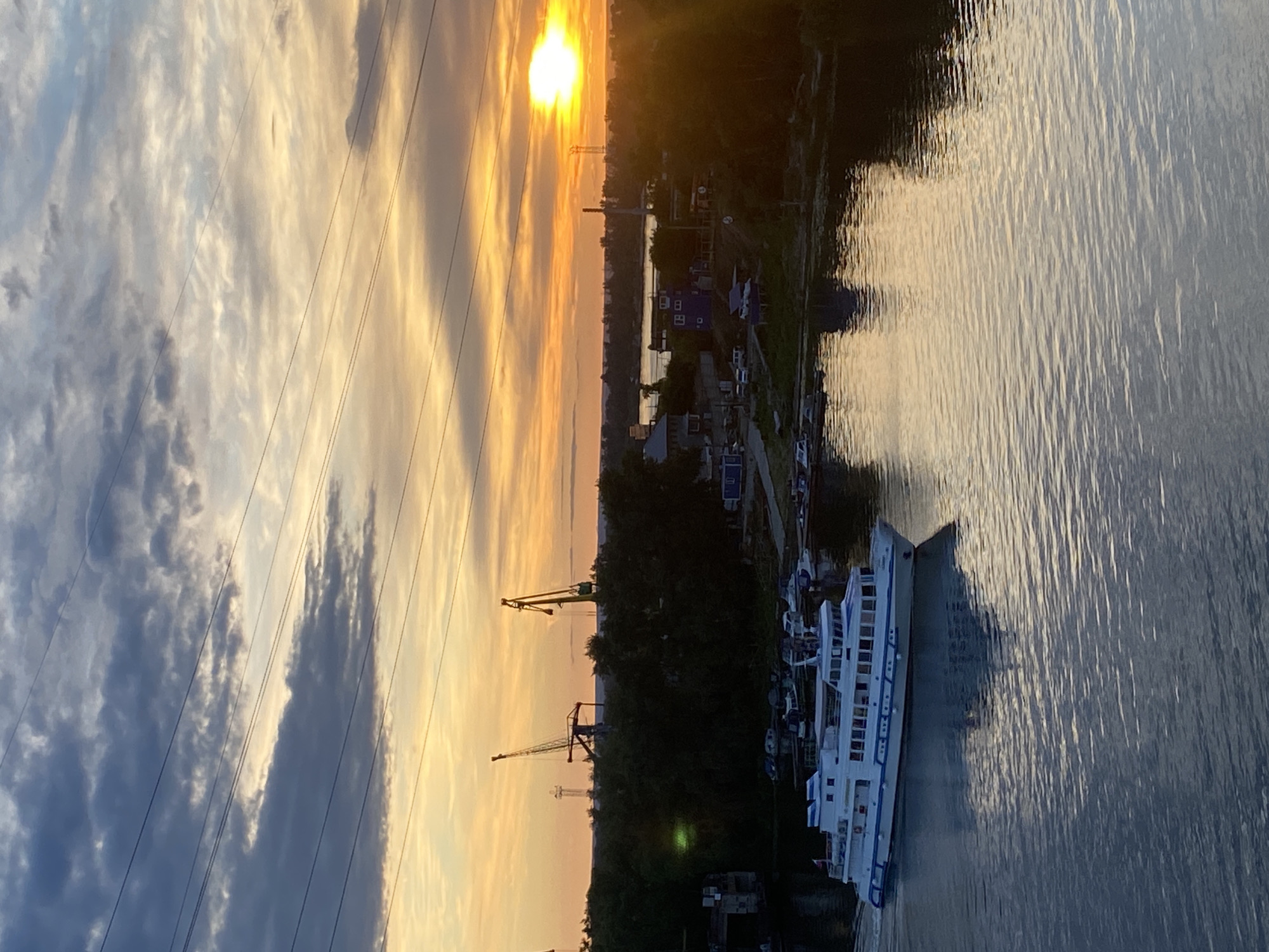 This image captures a sunset over a riverbank in Novokuybyshevsk, Russia. A white passenger boat with blue trim is traveling on the river in the foreground, leaving a wake. The river reflects the warm hues of the setting sun and the sky. On the far bank, a dense line of trees obscures much of the landscape, but glimpses of industrial structures, including two large cranes and some smaller buildings, are visible through the foliage. The sky is filled with dramatic clouds, painted with shades of orange, yellow, and grey, as the sun shines brightly on the horizon. Several power lines stretch across the top of the frame, silhouetted against the colorful sky. A few smaller boats are moored along the riverbank in the middle ground. The overall scene depicts a peaceful, industrial waterfront at dusk.