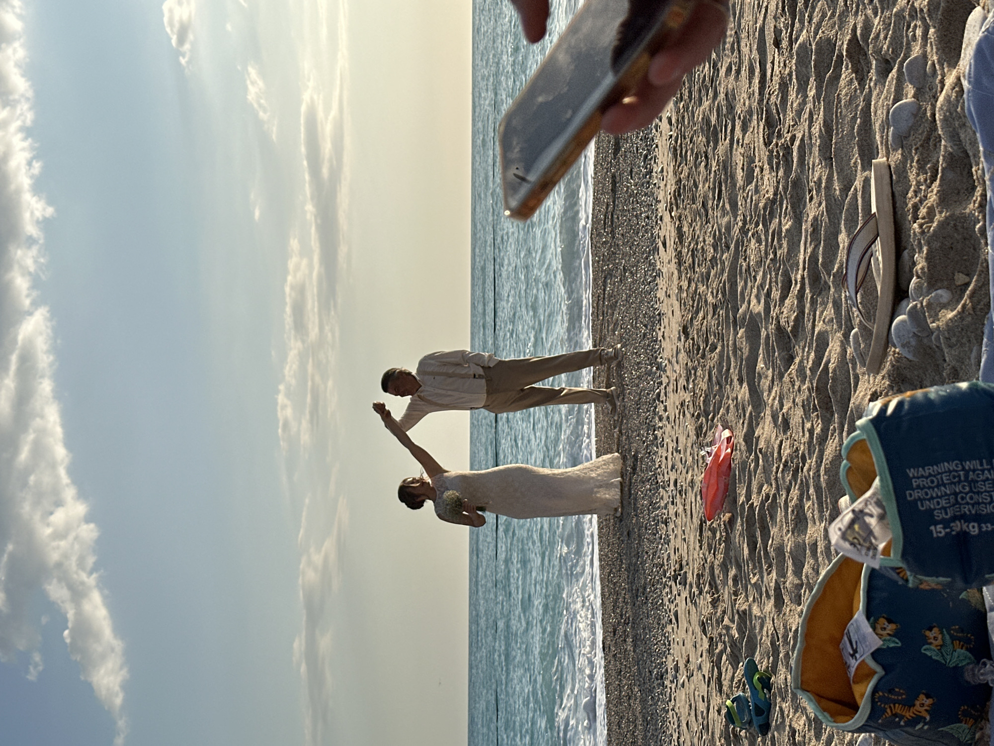 A wedding photo is taken on a beach in Fethiye, Turkey. The beach is a mix of sand and pebbles, with gentle waves washing ashore. A bride in a white lace dress and a groom in a light-colored shirt and khaki pants are positioned mid-frame, apparently dancing or posing. An individual is holding a phone, presumably to take the picture. To the right of the couple are items on the sand, including a pair of flip-flops, a child's inflatable swim vest, a red object, and blue sandals. The sky is blue with scattered clouds. The focus appears to be on the wedding couple.