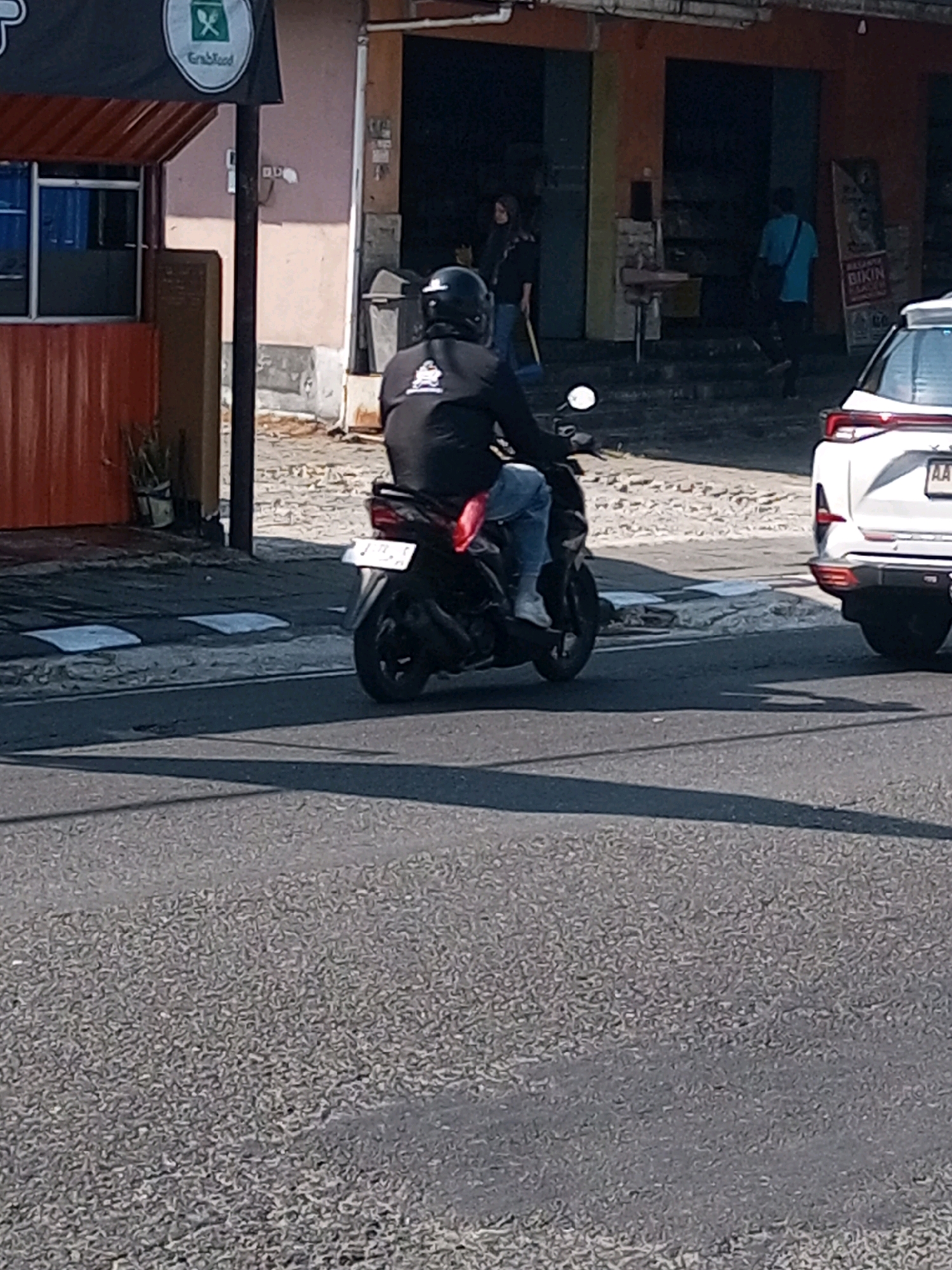 A person is riding a scooter on a street in Wonosobo, Indonesia. The scooter is black and the rider is wearing a black helmet and a black long-sleeved shirt with a logo on the back. They are also wearing blue jeans and white shoes. The scooter has a white license plate. In the background, there is a white car, a building with orange and brown accents, and a sign with "GrabFood" written on it. The street is paved with asphalt and there are some drainage covers visible. The overall lighting suggests it is daytime.