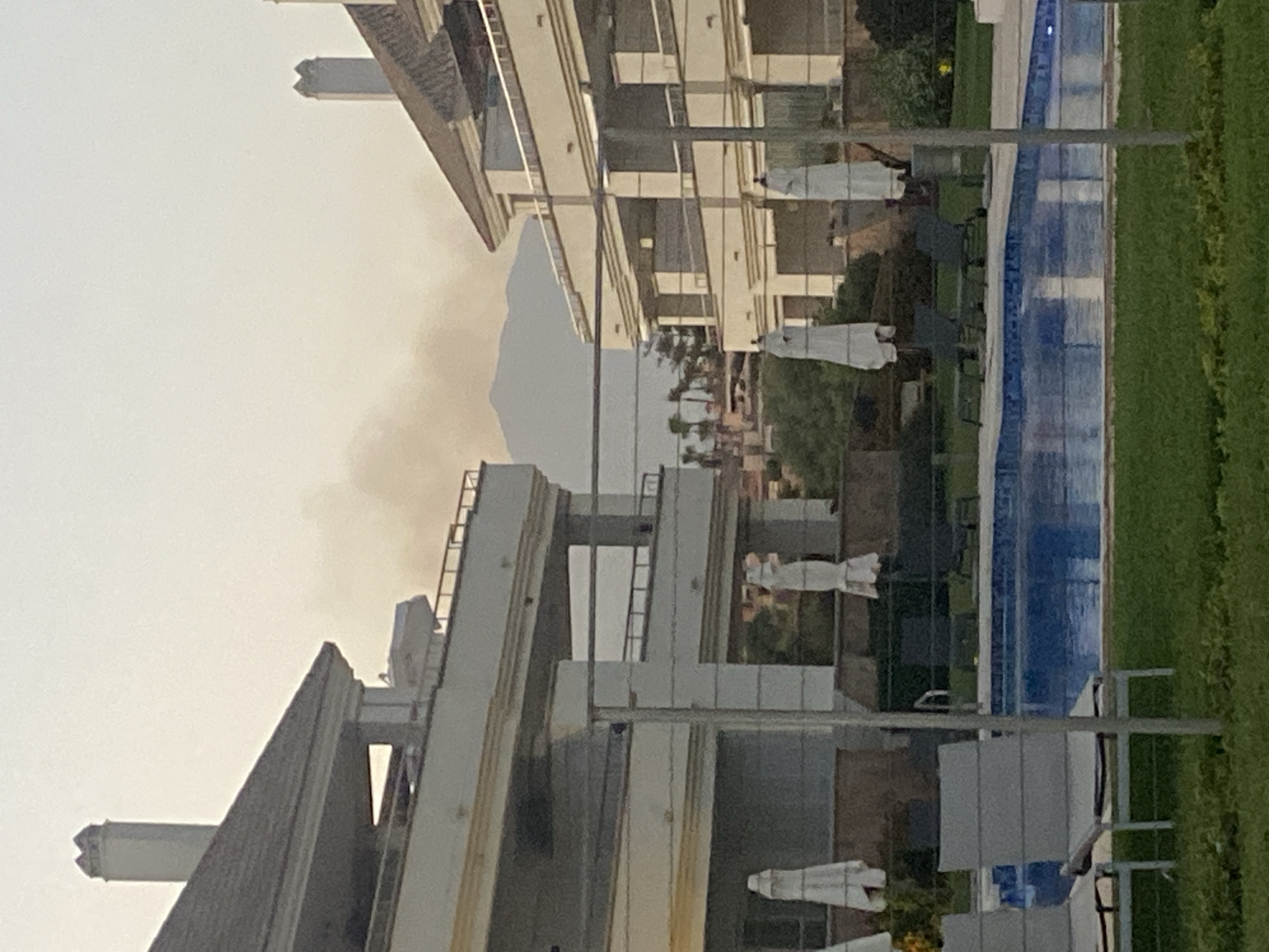 The image shows a resort-like setting with modern architecture and a swimming pool. In the background, there is smoke rising from a distant mountainous area, indicating a possible fire. The foreground features a swimming pool surrounded by manicured lawns and lounge chairs. Two white umbrellas are visible near the pool. The architecture suggests a hotel or condominium complex with balconies and decorative elements. The sky is overcast, with a warm, hazy tone. The location is identified as Playa Del Sol-Villacana, Spain, which is a coastal resort area.