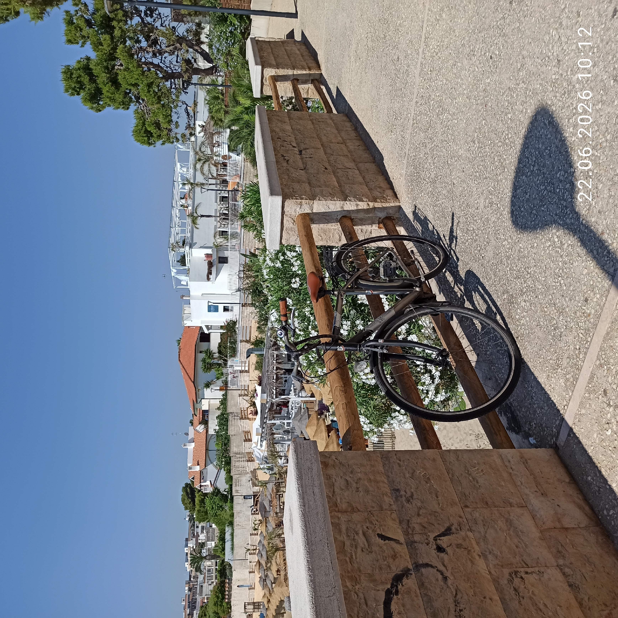 This image captures a scene in Trani, Italy, likely on a sunny day, as indicated by the clear blue sky and strong shadows. The primary objects in the foreground are a bicycle leaning against a wooden structure and a stone wall. The bicycle is dark-colored with black tires and a brown seat and handlebars, suggesting it's a functional object for transportation or recreation. The stone wall is textured and appears to be part of a pathway or retaining wall.
In the background, a vista of white buildings with red-tiled roofs unfolds, interspersed with lush green trees and palm trees. Some of these buildings appear to be part of a resort or residential area, with balconies and awnings visible. The presence of umbrellas and what looks like a beach area in the mid-ground suggests a coastal or seaside location.
The time of day is indicated by the timestamp "22.06.2026 10:12" visible on the right side of the image, suggesting it was taken in the morning. The shadow cast by an unseen object, possibly a lamppost or a tree, is prominent on the paved ground, further emphasizing the sunny weather. There are no people visible in the frame, creating a sense of quiet or perhaps a moment before activity begins. The overall impression is a peaceful, sun-drenched Mediterranean scene.