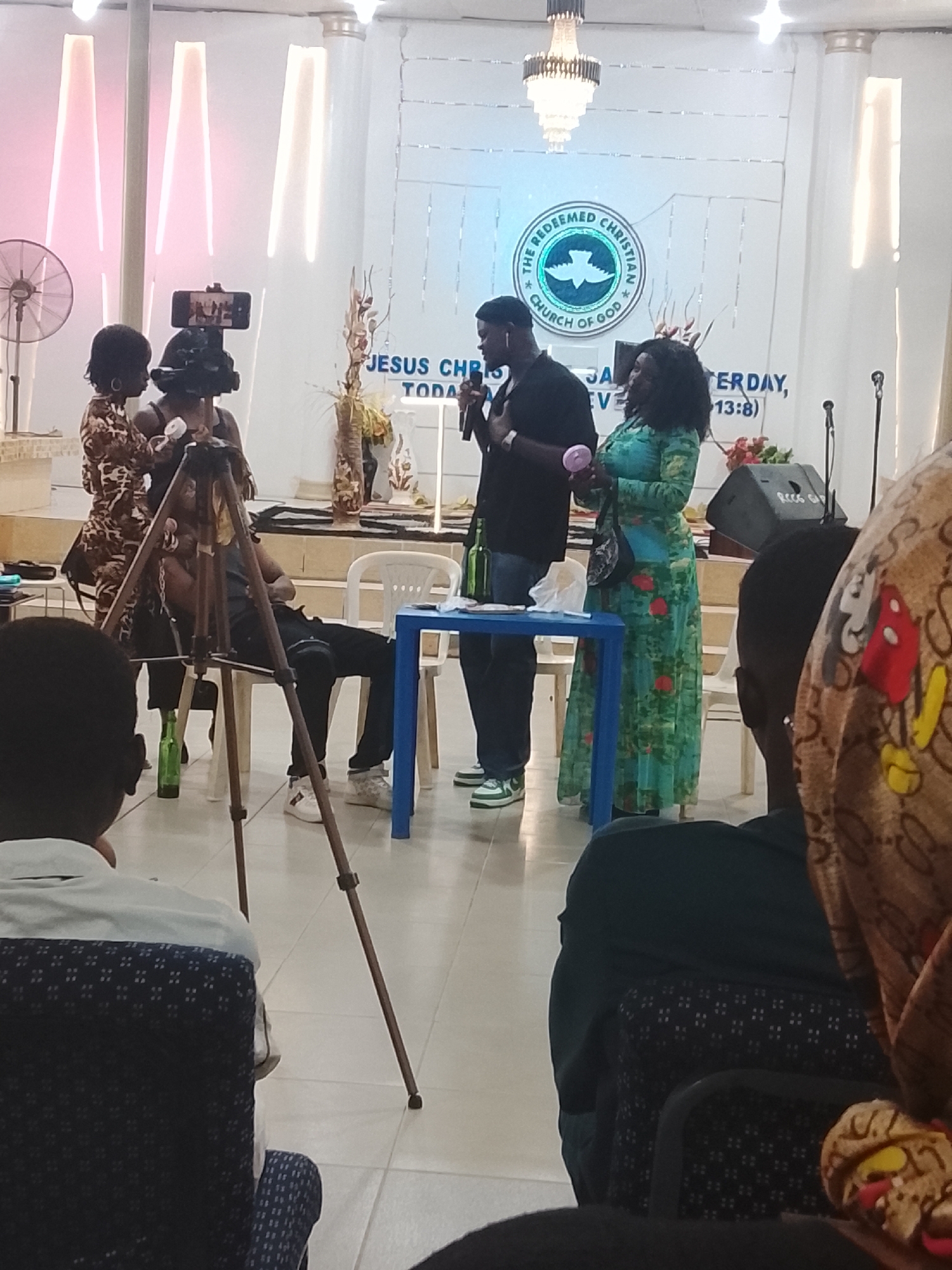 A medium indoor shot shows a man speaking into a microphone on a small stage, while a woman in a long patterned dress stands next to him holding a pink object. Two women are to the left, in front of a tripod on which a smartphone is attached. A man is seated on a chair behind the tripod. A blue table stands between the speaker and the woman in the dress. The backdrop of the stage features the logo of "The Redeemed Christian Church of God". The scene appears to be recorded or broadcast, while other individuals are seated in the foreground, presumably as audience members. The event is happening in Jos, Nigeria.