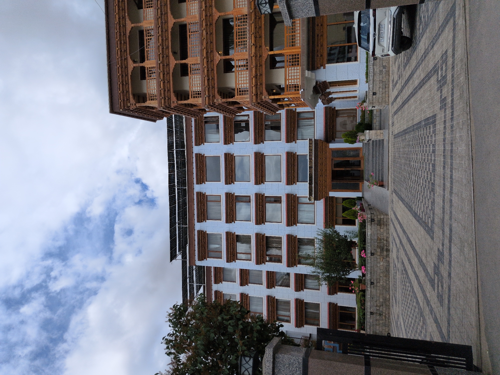 The image shows a multi-story hotel building with a traditional architectural style. The building has a light blue facade and is adorned with intricately carved wooden balconies on the right side. The roof of the building on the left has solar panels installed. In front of the hotel, there is a paved driveway with a decorative pattern. A white SUV is parked on the right side of the driveway, facing away from the camera. The entrance to the hotel features steps leading up to double wooden doors. There are trees and flowering plants in planters in front of the building. The sky is partly cloudy with a mix of blue sky and white clouds. The scene is likely in Leh, India, given the architectural style and the license plate of the car, which appears to be from the region. No people are visible in the image.