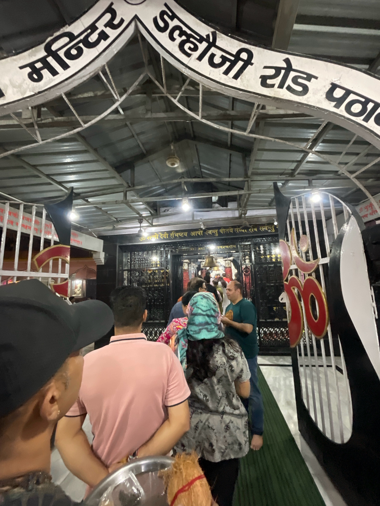 The image shows the entrance to a temple in Pathankot, India. The entrance archway has text in Hindi, which appears to say "Mandir" and "Dalhousie Road Pathankot". A crowd of people are entering or exiting the temple. In the foreground, a person wearing a black baseball cap is partially visible from behind. Behind them, a person in a pink polo shirt is facing away from the camera, with their hands clasped behind their back. To the right, a metal gate with decorative red and black elements is visible. The temple interior appears to have a dark facade with metal railings and a bell hanging. Several lights illuminate the area. The ground is covered with a green carpet.
