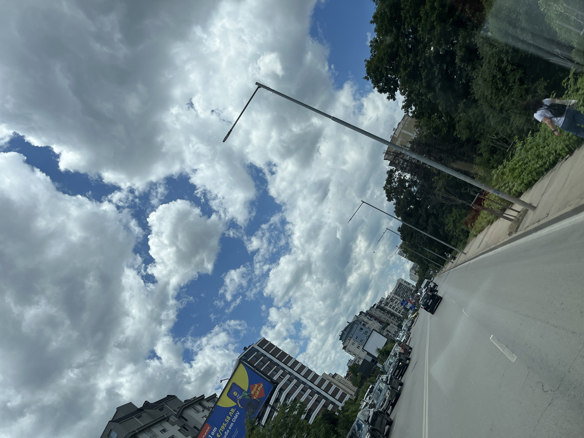 A low-angle shot captures a city street from a vehicle, looking forward and slightly upwards. The sky is filled with dramatic, heavy white and gray clouds, with patches of blue visible. A tall, modern building with a large billboard is on the left side, advertising what appears to be a sale for Lidl Plus. Several cars are driving on the multi-lane road, which has white lane markings. On the right side of the street, there are dense green trees and vegetation lining a sidewalk. A streetlamp is visible on the right, extending diagonally into the sky. A person wearing a white shirt and blue jeans, with dark curly hair and a backpack, is walking on the sidewalk.