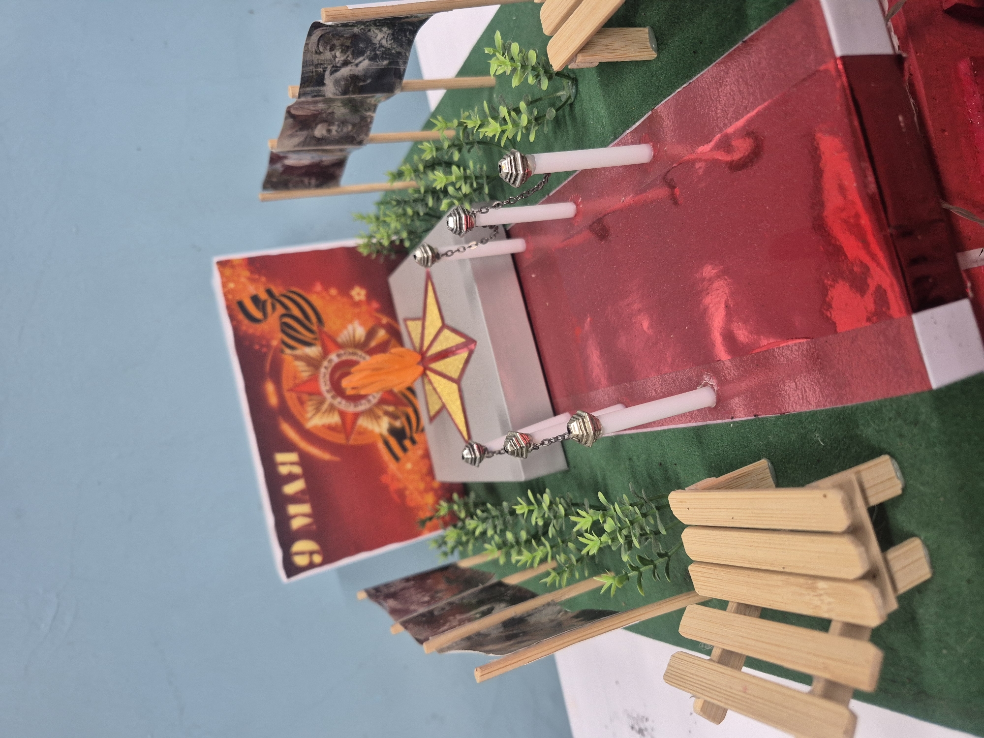 The image depicts a miniature model of a memorial or monument. The model includes a red walkway, a central pedestal with a five-pointed star, symbolic eternal flame and miniature flags with images of soldiers. Green foliage simulates landscaping around the monument. A poster in the background shows the Russian text "9 мая," meaning "May 9th," referencing Victory Day, which commemorates the Soviet Union's victory over Nazi Germany in World War II. Wooden benches are also included in the model. The model appears to be set against a blue background, which could be a table or wall. It is impossible to definitively identify the location of the memorial or monument in the city of Chaikovsky, Russia, based solely on this image.