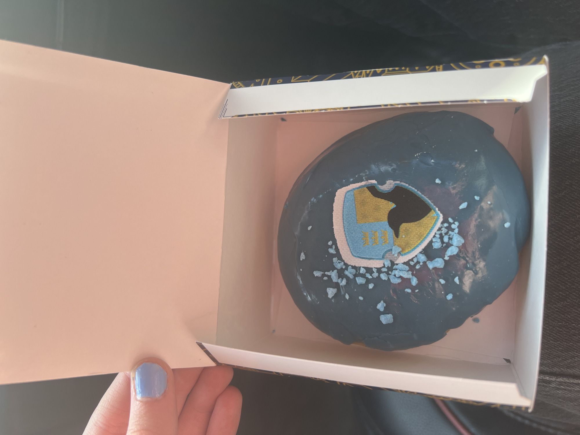 A close-up, overhead shot shows a single, blue-frosted donut in a white box. The donut has a crest-like decoration on top, featuring a black bird silhouette against a yellow and blue background, surrounded by scattered light blue sugar crystals. A hand with light blue nail polish is visible on the left side of the frame, holding open the lid of the box. The box has patterned sides with navy blue and gold geometric designs. The scene appears to be inside a vehicle, with dark upholstery visible in the background and the side of the box catching the light from outside, suggesting it might be daytime. The location in Revelstoke, Canada, is not discernible from the image.
