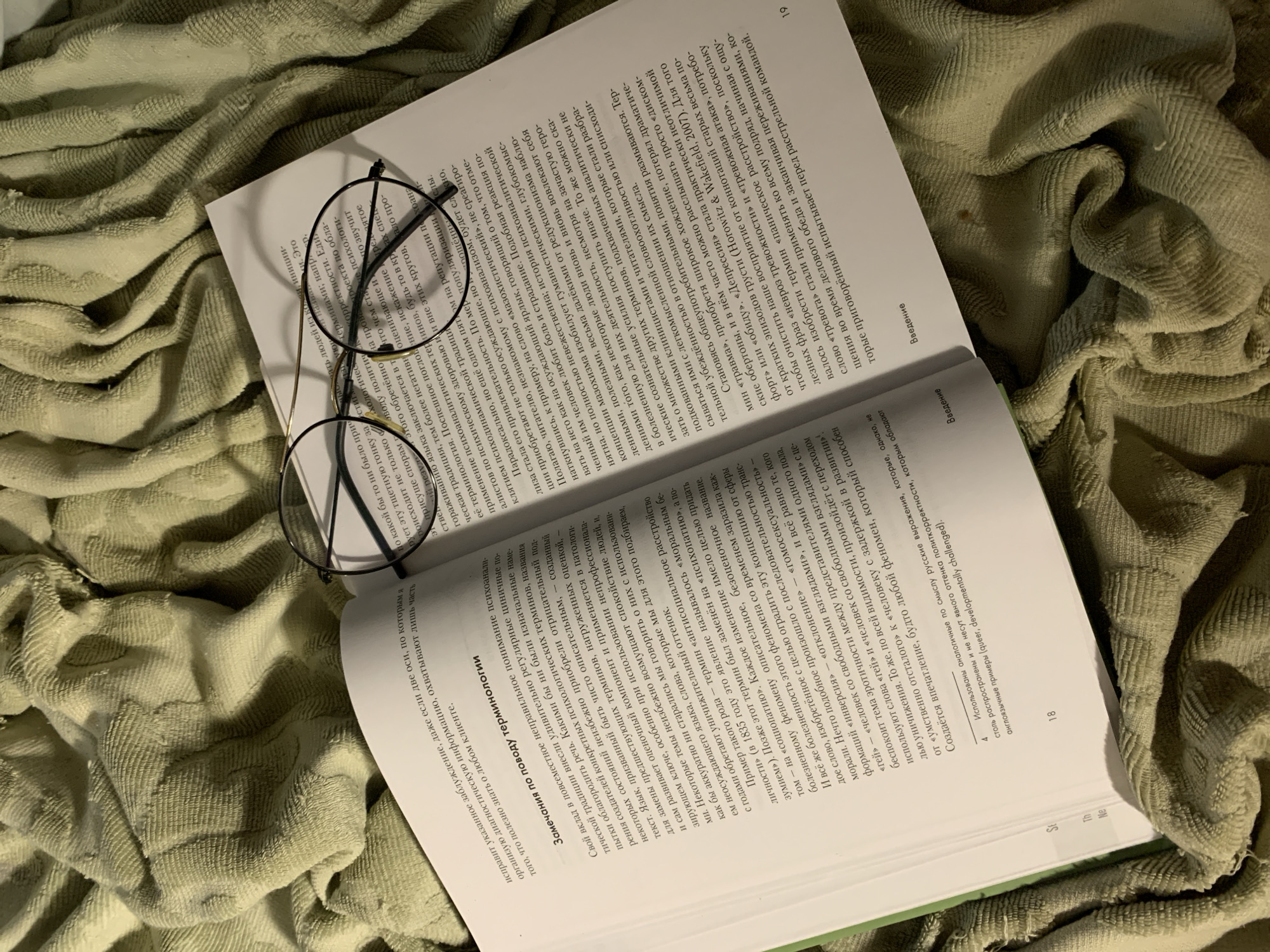 The image shows an open book with eyeglasses resting on its pages, lying on a crumpled, light green fabric. The book is positioned at an angle, with the bottom right corner of the book closest to the viewer. The pages are filled with Russian text. The eyeglasses have a thin, dark metal frame. The fabric underneath is rumpled with varying textures and shadows. The lighting is soft and diffused, creating a subtle contrast between the lighter areas of the book and the deeper folds in the fabric.