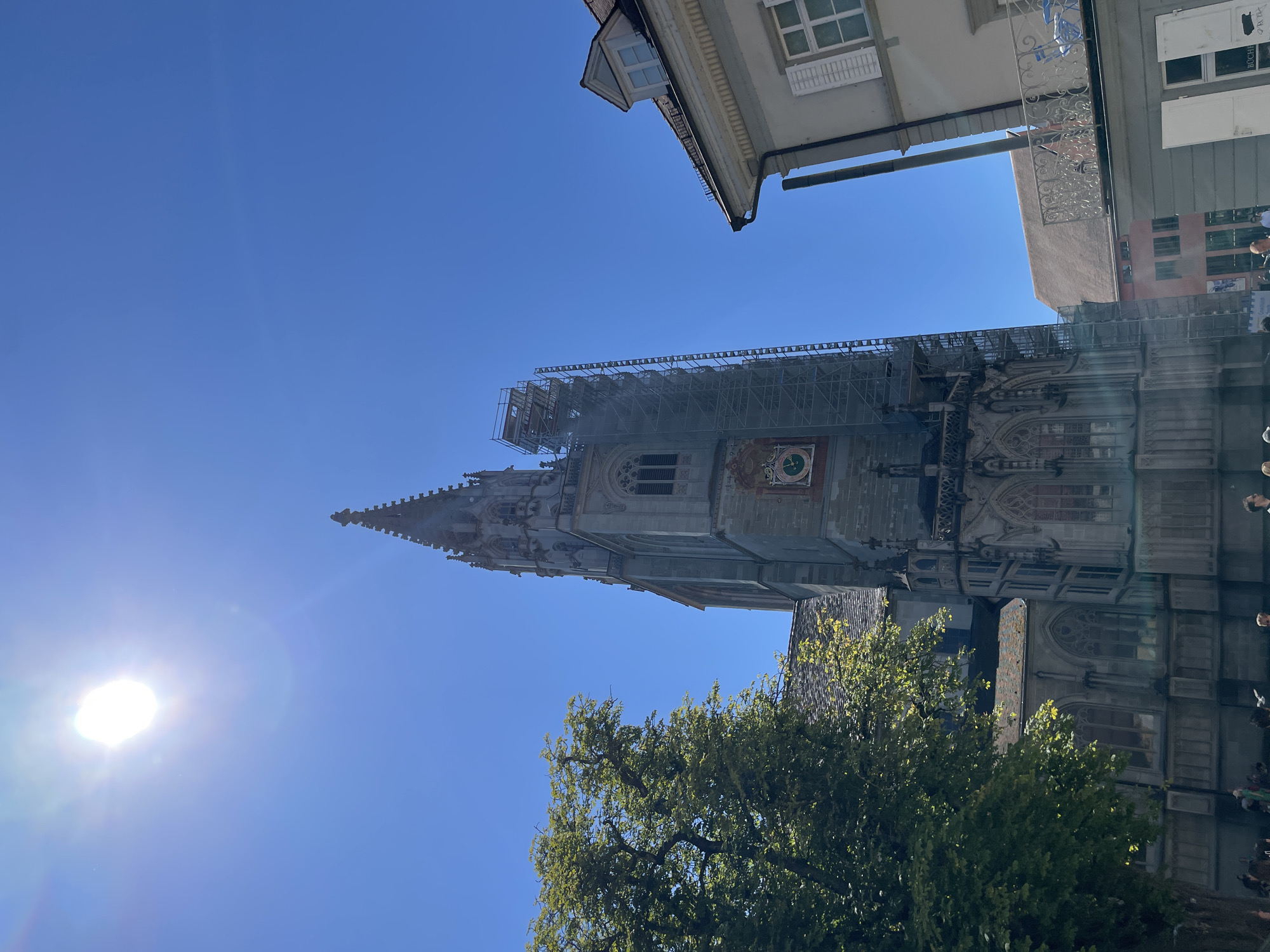 The image shows a bright, sunny day in Constance, Germany, with a clear blue sky. The focal point is the Constance Minster, a large Gothic cathedral. Its tall spire and intricate stone facade are visible, with scaffolding on one side, suggesting ongoing renovation or maintenance. The cathedral is surrounded by lush green trees. To the right of the Minster, a lighter-colored building with white shutters and a balcony is visible. The street level shows a few indistinct people walking.
