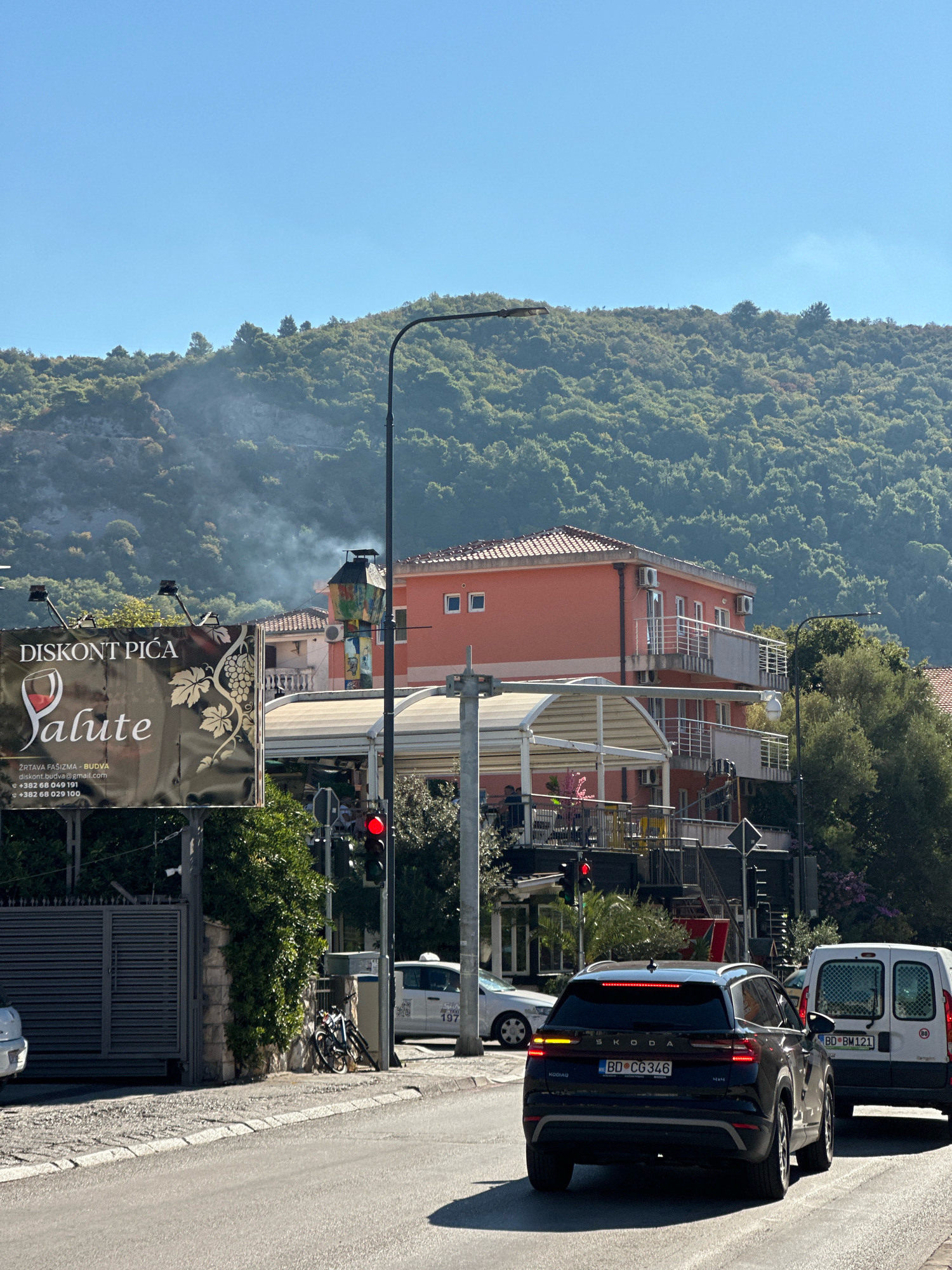 The image captures a street scene in Budva, Montenegro, on a clear day with a bright blue sky. In the foreground, a black Skoda Kodiaq SUV is driving away from the camera. To its right, a white van is also moving forward. The road is paved with asphalt and bordered by a cobblestone sidewalk. On the left side of the street, a large billboard advertising "DISKONT PIĆA SALUTE" is prominently displayed. Behind the billboard, a gray metal gate is visible, suggesting an entrance to a property. Adjacent to the gate, several bicycles are parked leaning against a stone wall.
Further back, a multi-story building with an orange facade and red-tiled roofs stands. It features several balconies with white railings and air conditioning units visible on the exterior. A white canopy extends over an outdoor seating area on the lower level of this building. A street lamp with a visible light fixture stands on the right side of the road. A traffic light displaying a red signal is positioned near the intersection. In the background, a lush, green, tree-covered hillside rises, with a faint wisp of smoke emanating from its slopes, possibly from a chimney. A white taxi with the number "197" visible on its roof is parked on the right side of the road, behind the bicycles. The overall environment suggests a sunny day in a coastal town.