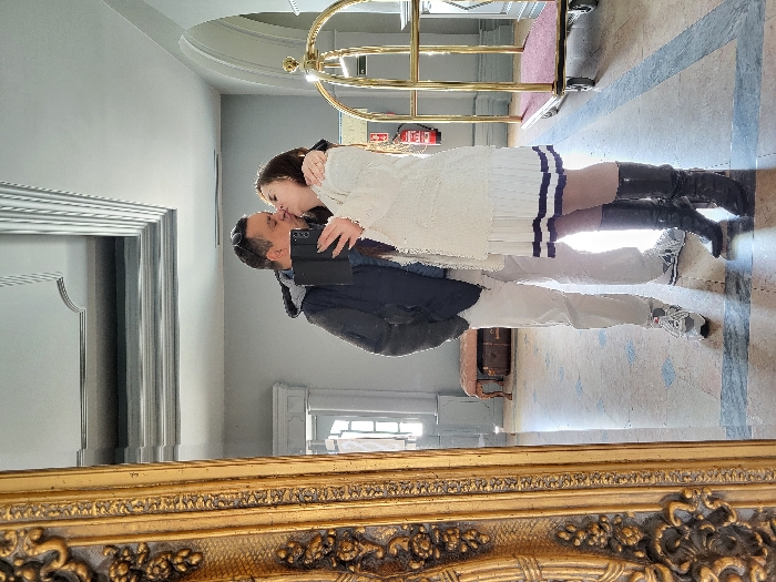 The image depicts a mirror selfie of two individuals kissing in what appears to be a well-appointed interior space, possibly a hotel lobby, located in São Pedro do Sul, Portugal. A male, wearing a dark jacket, light trousers, and athletic shoes, is on the left, embracing a female on the right. The female wears a light-colored top, a white pleated skirt with dark stripes, and black high-heeled boots. She holds a dark foldable smartphone in her right hand, capturing the reflection. Both individuals are engaged in a kiss.
The environment features a large, ornate gold-framed mirror, partially visible on the left. The floor is light-colored tile with darker accent patterns. In the background, there is a light grey wall with architectural molding, an archway on the right, and a window or glass door on the left providing natural light. A small, upholstered pink bench with a dark trunk underneath is visible in the mid-left background. A red fire extinguisher is mounted on the wall further back, and a brass-colored, two-tiered luggage cart with a red carpeted base is positioned on the far right. The ceiling is light-colored with decorative recesses and molding.