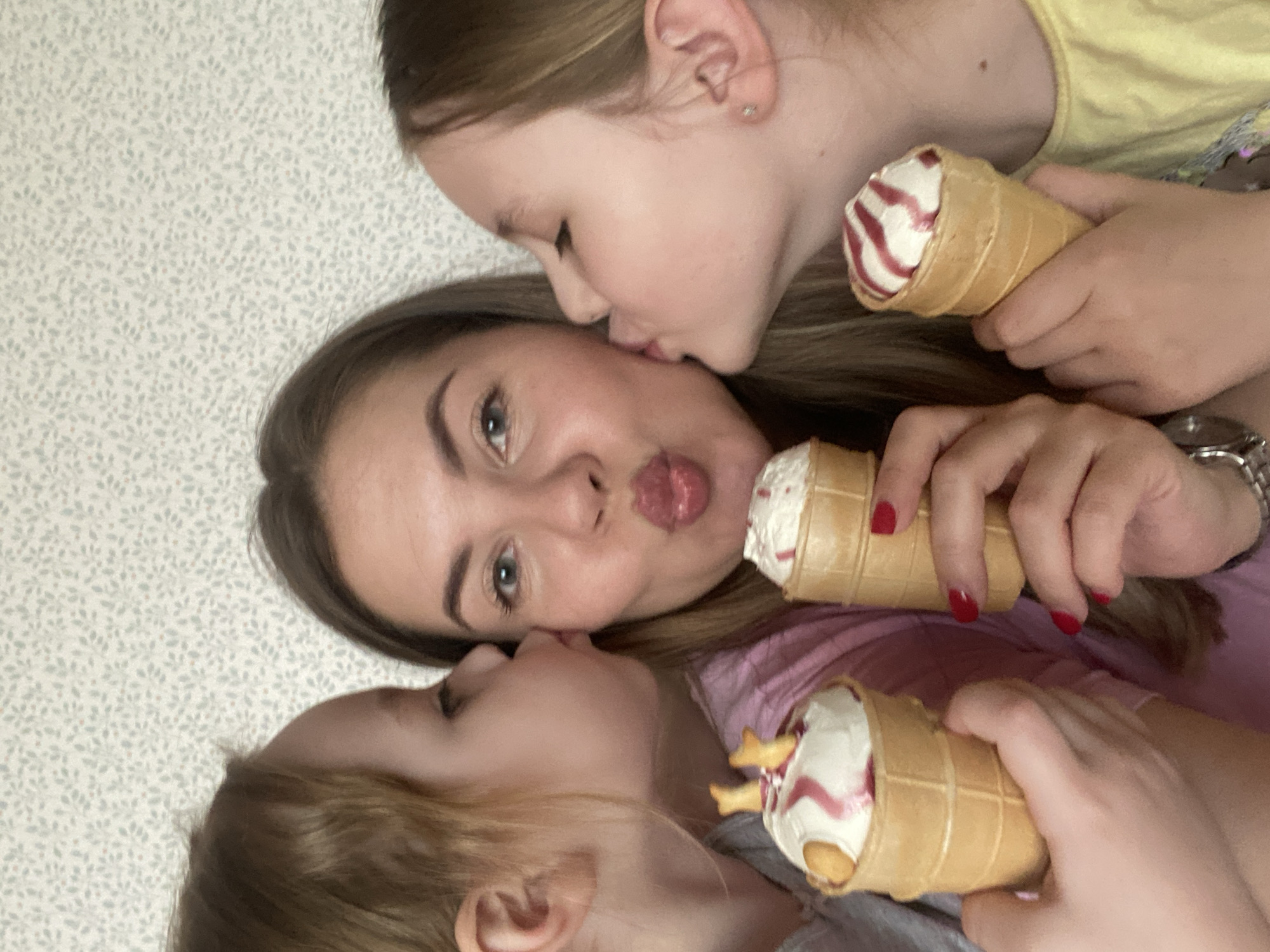 The image depicts a selfie of a young woman with two young girls, all holding ice cream cones. The woman is positioned in the center with her face tilted towards the camera, puckering her lips. A girl is on either side of her, each giving her a kiss on the cheek. Each person is holding an ice cream cone. The background is a wall with a floral wallpaper design. The location is unknown and cannot be determined from the image.