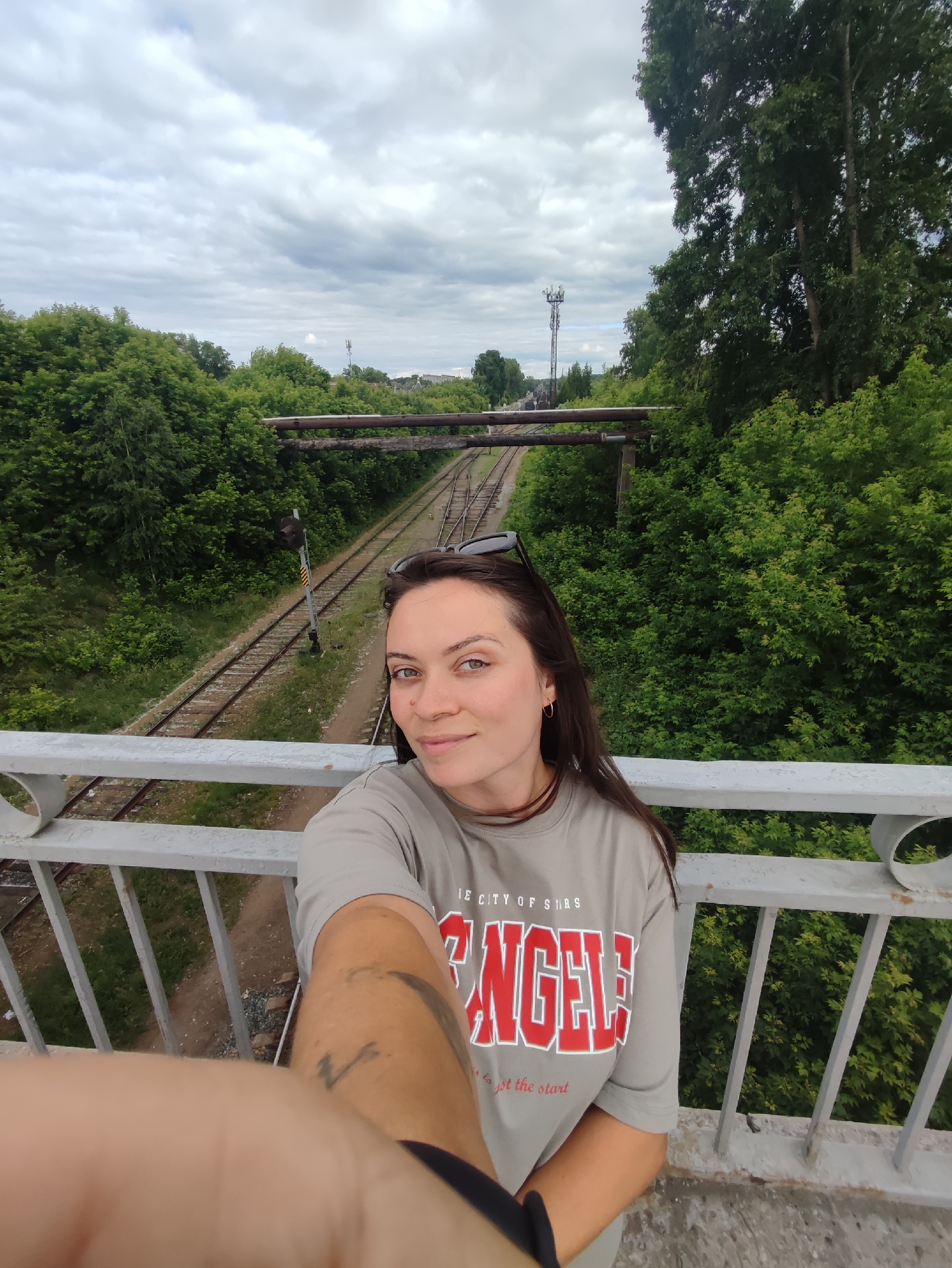 In this candid selfie, a young woman with dark brown hair, wearing a grey t-shirt with red lettering that reads "ANGELS", stands on a pedestrian overpass. Her sunglasses are perched on her head, and she has a light expression. The overpass, made of grey concrete and metal railings, overlooks a railway track that curves into the distance. The track is surrounded by lush green trees and foliage. In the background, a tall telecommunications tower is visible against a cloudy sky. The scene appears to be located in Chaikovsky, Russia, given the context of the railway and the natural landscape.