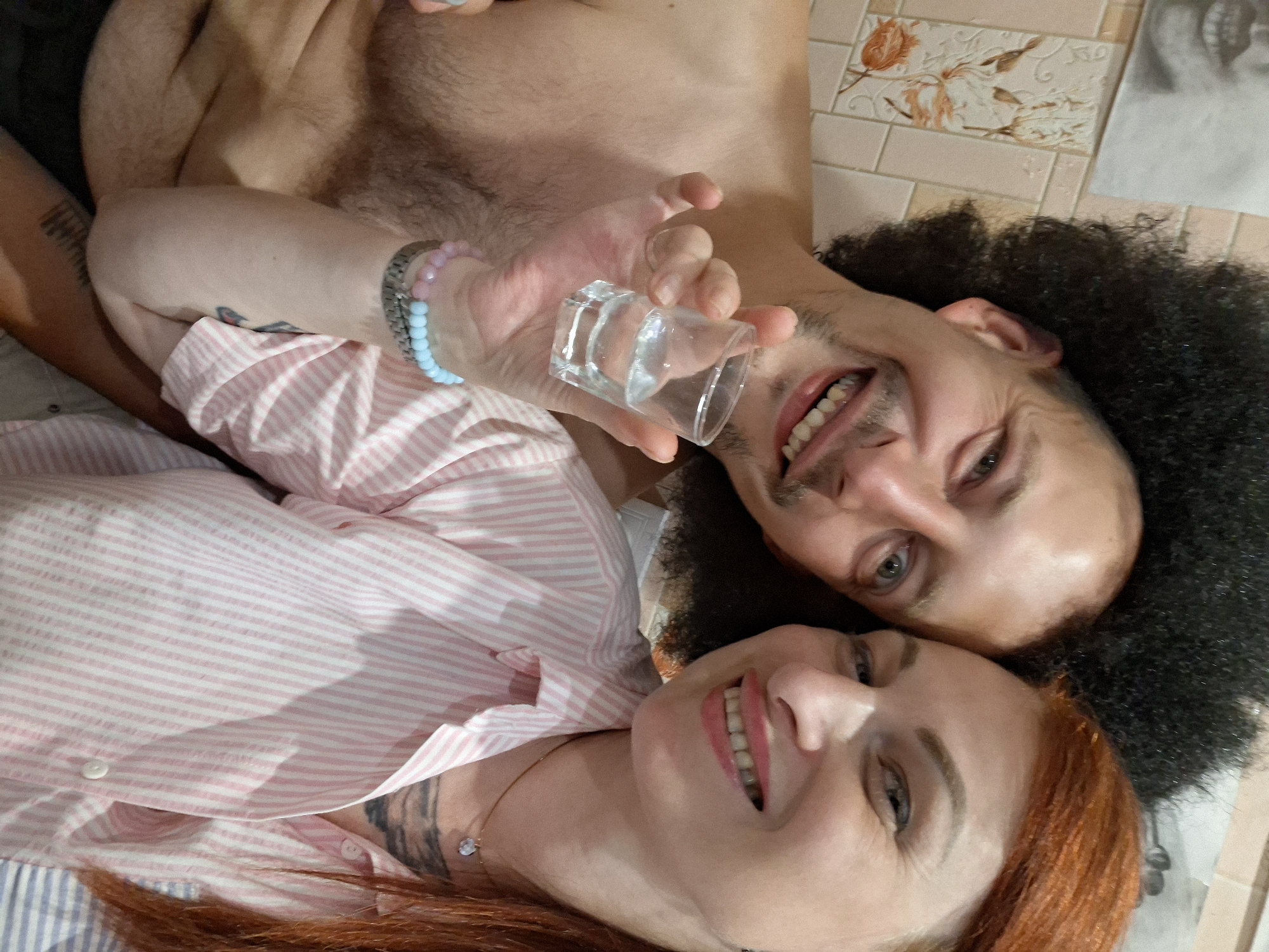 In the foreground of this image, a man and a woman are close together, appearing to be taking a selfie. The man, on the left, has a large afro hairstyle and a wide smile, holding up a shot glass filled with a clear liquid. He is shirtless, revealing his chest and shoulder. The woman, on the right, has long red hair and is smiling broadly. She is wearing a pink and white striped shirt. The background appears to be a bathroom with tiled walls. No specific location within Myadzyel, Belarus can be determined from the visual information provided.