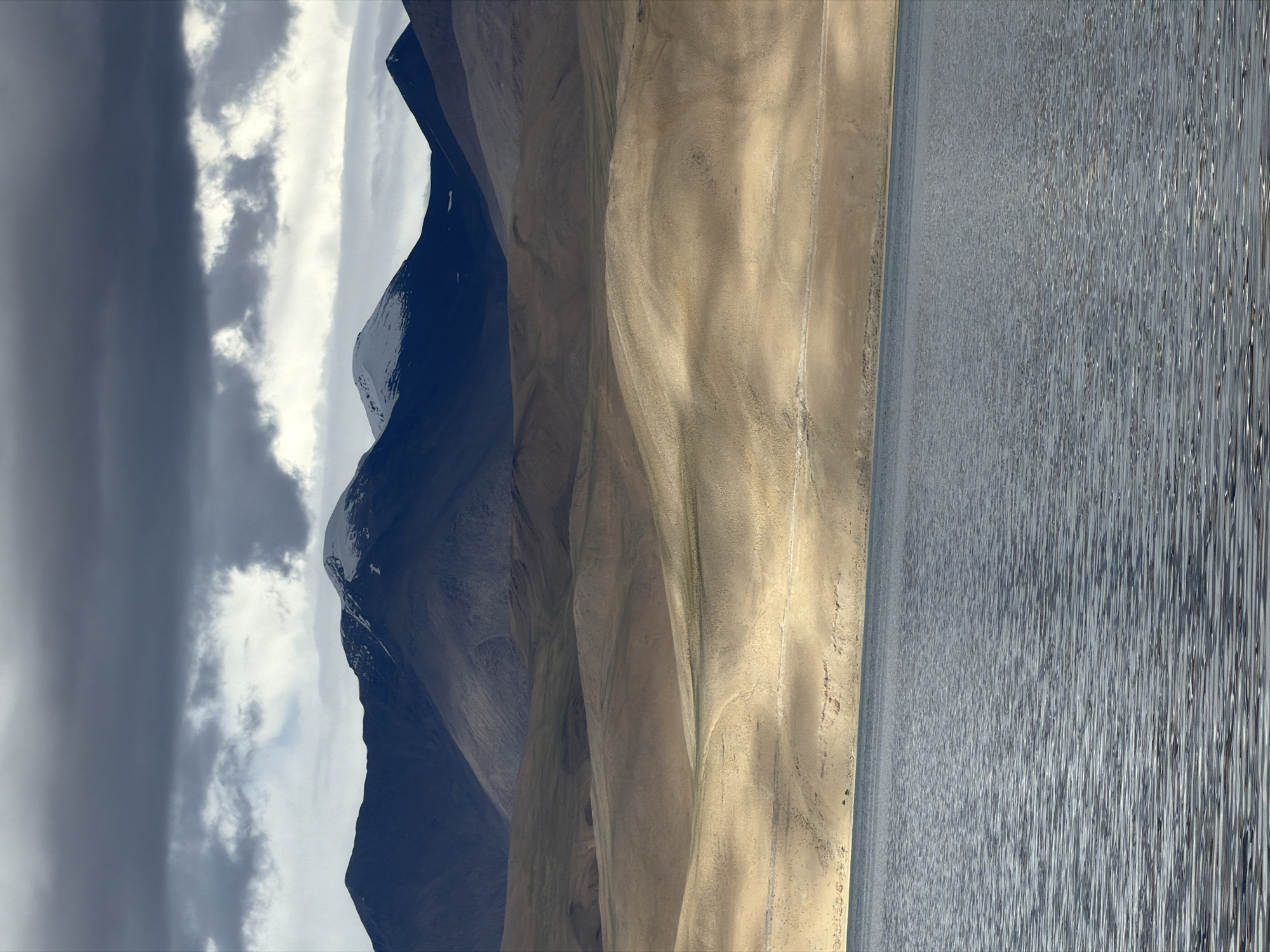 The image depicts a panoramic landscape of a mountainous region with a lake in the foreground. In the background, dark, heavy clouds hang over sharply defined mountain peaks, some of which are capped with snow. Below the highest peaks, rolling hills with muted earth tones stretch towards the foreground. The middle ground features a series of undulating, sandy slopes illuminated by sunlight, creating distinct shadows and highlights. A narrow, faint track or road is visible traversing these slopes. The foreground is dominated by a calm body of water, likely a lake, with a rippled surface reflecting the muted light. The overall scene suggests a remote, high-altitude environment, possibly in Ladakh, India, characterized by dramatic geological formations and atmospheric conditions. No people or identifiable man-made structures are prominent in the scene, aside from the faint track.