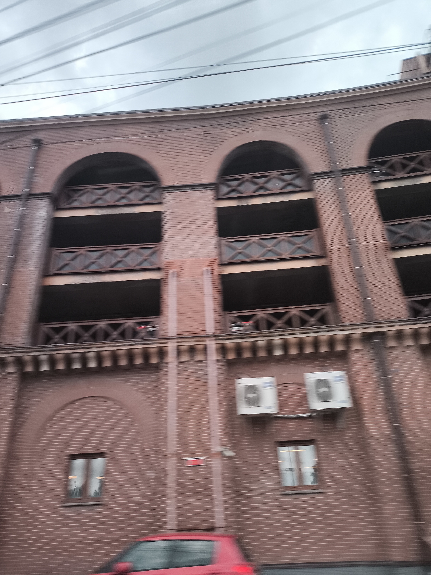 The image, captured in motion, shows a multi-level brick building with arched openings on the upper floors, each with wooden lattice railings. Two air conditioning units are mounted on the brick wall of the lower level. Below these, a small, dark-framed window is visible, reflecting the sky and perhaps some interior lighting. A red car is partially visible in the foreground, also in motion, suggesting the photograph was taken from a vehicle. The sky above is overcast and appears to have multiple power lines running across it. The overall impression is of an urban environment, likely a parking garage or a similar structure, viewed from street level.