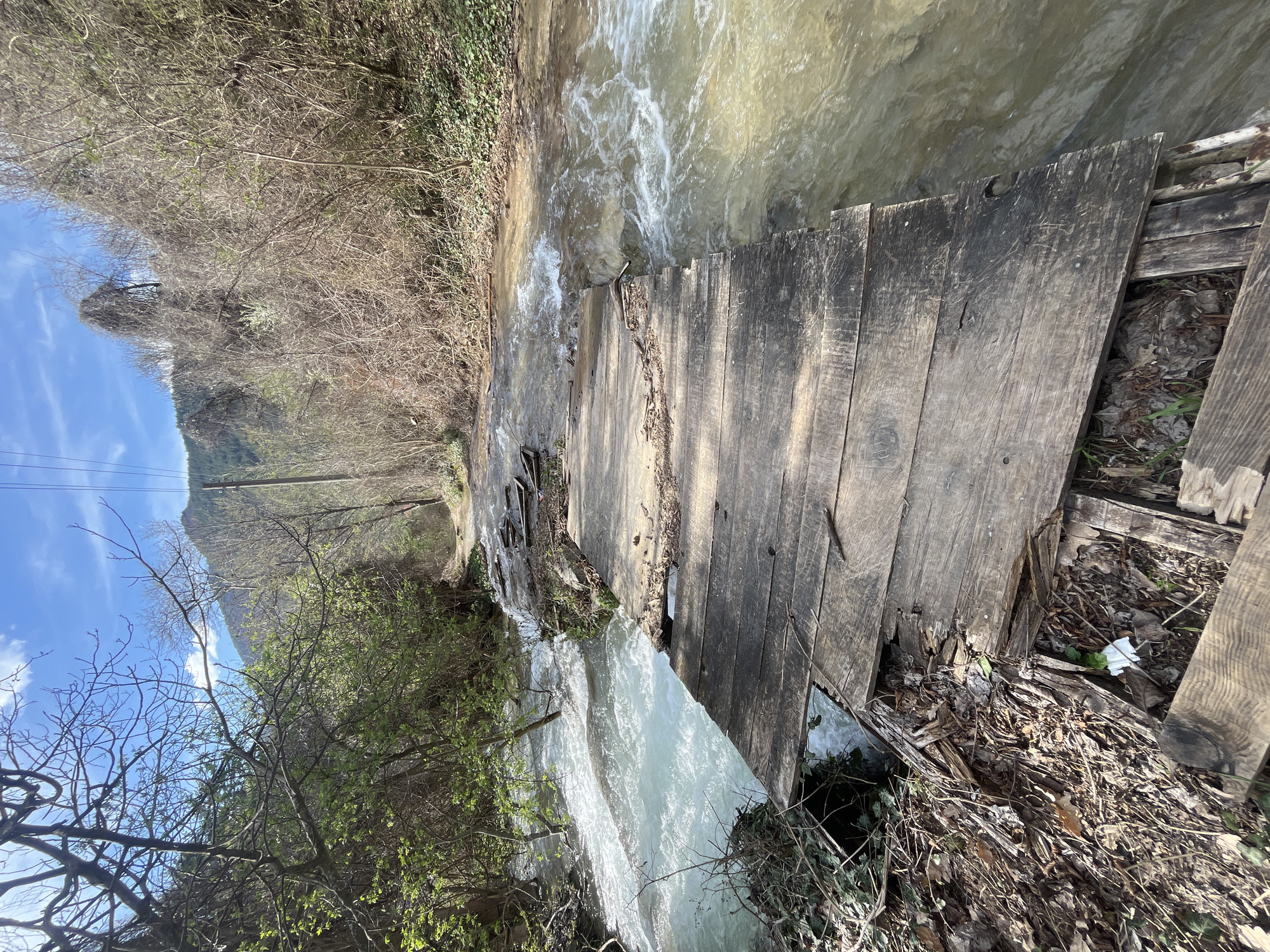 The image depicts a dilapidated wooden footbridge spanning a fast-flowing stream. The bridge is constructed of weathered planks, some of which are broken or missing, revealing the stream beneath. Debris and vegetation are visible both on the bridge and along the banks of the stream. The stream itself is turbid, with white water indicating strong current. The surrounding environment consists of foliage, including both trees with budding leaves and bare branches, and a mountain in the background. A blue sky is visible with some light clouds. Based on information given, this is located in Bachkovo, Bulgaria.