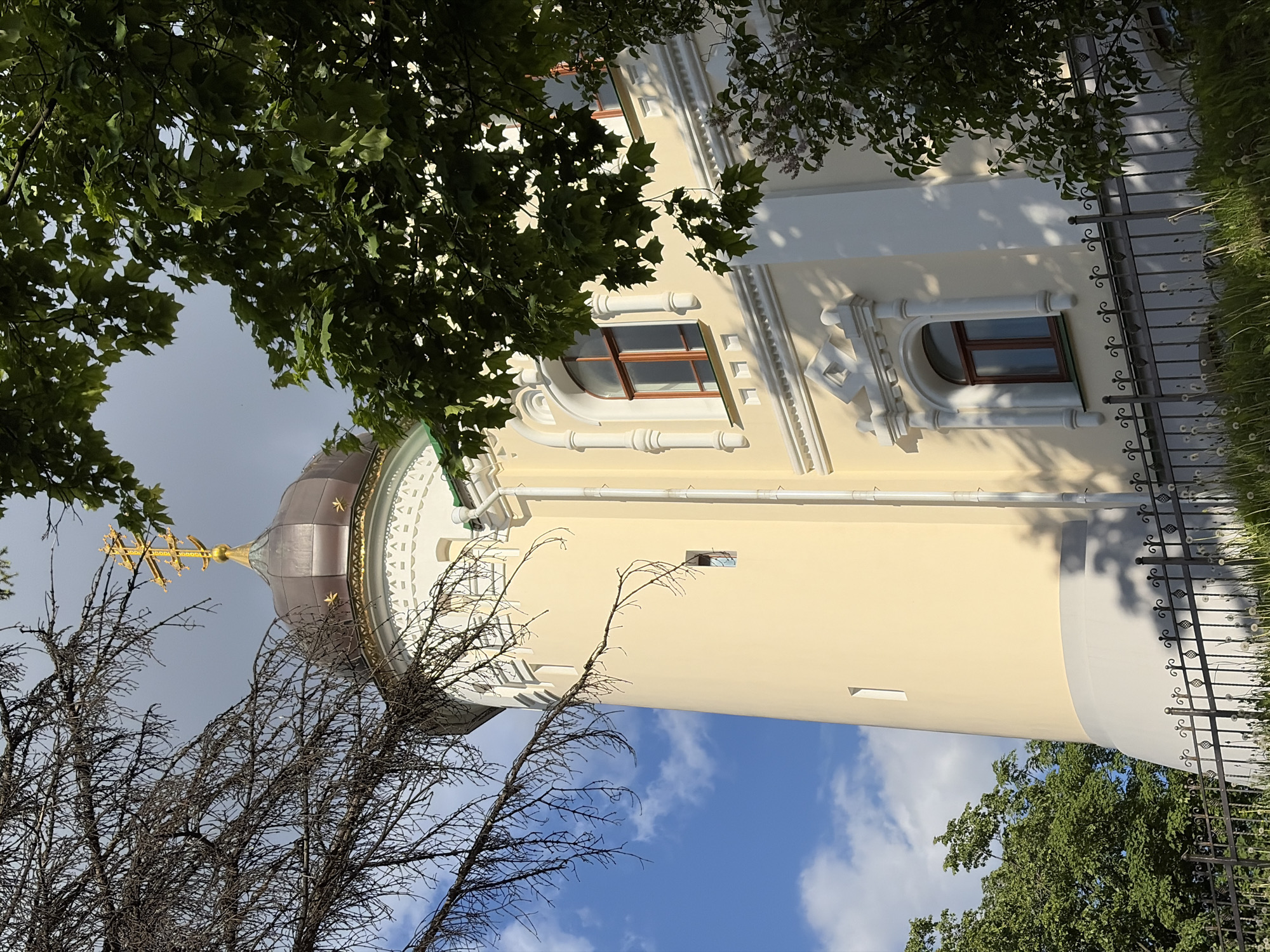The image captures a building, possibly a church, with a golden dome and a cross on top, partially obscured by bare tree branches in the foreground. The building's façade is light yellow, with white architectural details and windows with brown frames. Green trees are visible on both sides, framing the structure. A metal fence runs along the side of the building. The sky is blue with scattered white clouds. The location is Minsk, Belarus.