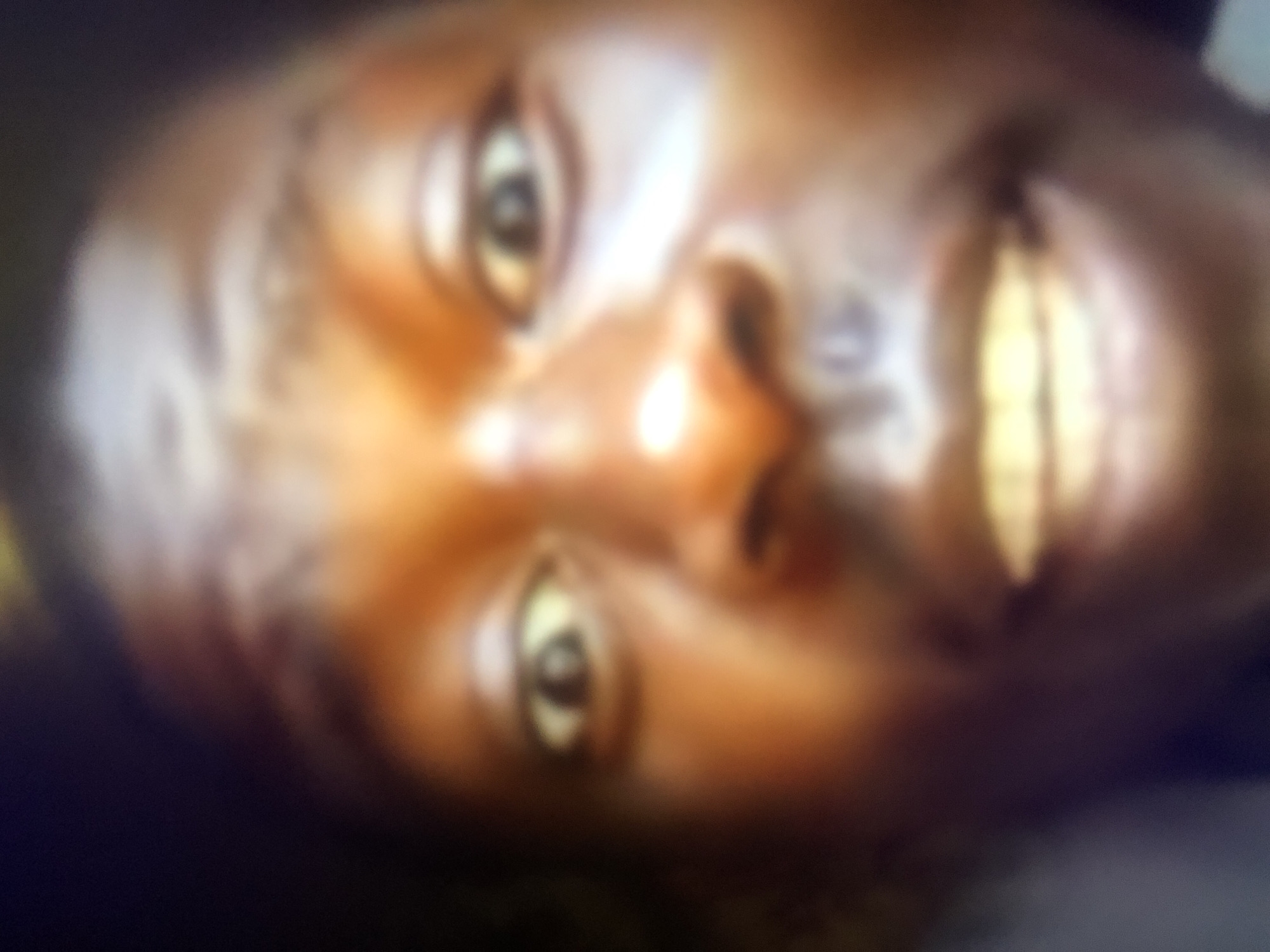 The image is a very blurry, close-up, frontal shot of a person's face, likely taken with a phone camera. The subject has dark skin and is looking directly at the camera. The lighting is uneven, with bright highlights on the forehead and cheeks, and darker areas on the left side of the face and in the background. The person's mouth is open, revealing teeth. The image quality is poor, with a lot of noise and lack of detail, making it difficult to discern specific features. No specific location in Howayi, Nigeria, can be identified from the image itself.