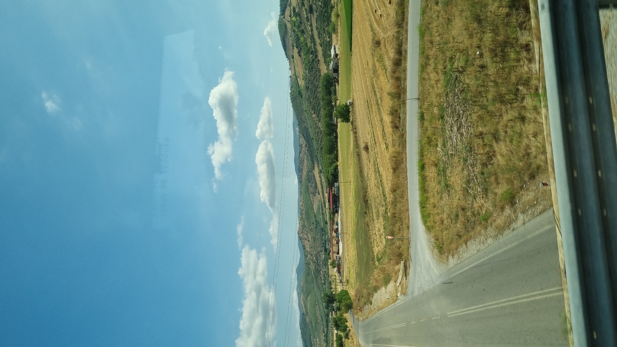 The image captures a rural landscape on a clear day, dominated by a vast blue sky with scattered white clouds. A winding asphalt road, marked with double yellow lines, leads into the distance from the lower left of the frame. The road curves to the right, bordered by dry, golden-brown vegetation and scattered green shrubs.
In the mid-ground, a flat, agricultural field with harvested crops, appearing as golden stubble, stretches across the scene. Beyond the field, a cluster of buildings with red roofs, possibly a small commercial establishment or farm, is visible. A line of trees separates the field from a gently rolling hillside covered in green vegetation and olive groves, typical of the Thessaly region. In the far distance, more hills create a layered horizon.
A metal guardrail runs along the bottom of the frame, suggesting the photograph was taken from a vehicle or elevated position. There are no visible people or animals in the image. The overall impression is one of a serene, rural setting bathed in sunlight. The specific location in the city of Trikala, Greece, cannot be definitively determined from the visual information alone, though the landscape is consistent with rural areas surrounding the city.