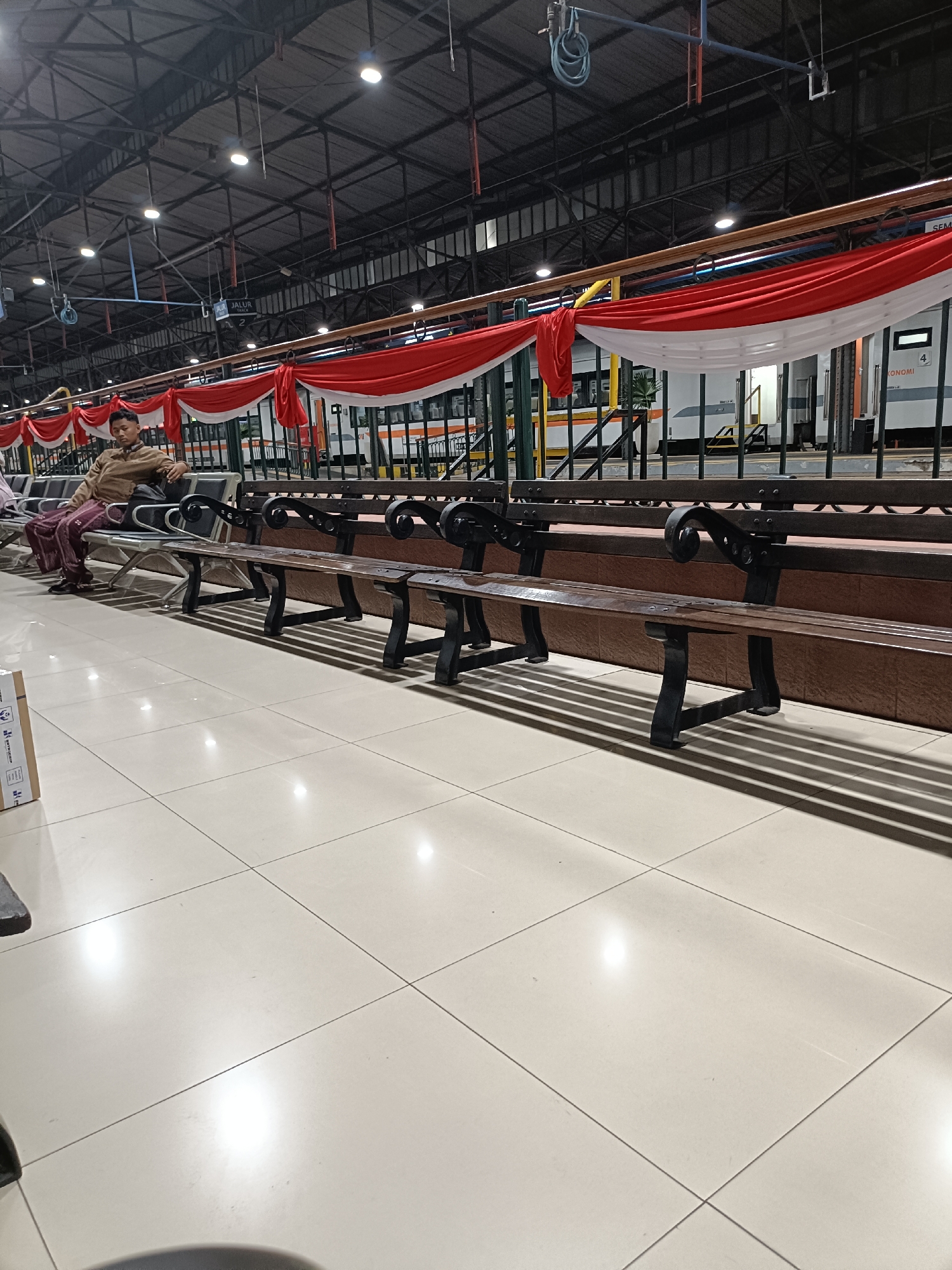 The image depicts an indoor train station waiting area, likely in Semarang, Indonesia, based on the provided context of the image. A young man, wearing a light brown jacket and patterned trousers, sits on a metal and wooden bench. He is looking to his left, with a bag resting on the bench beside him. The waiting area features rows of similar benches facing a train platform. Red and white fabric banners are draped along a railing above the platform, suggesting a festive or patriotic decoration. The floor is tiled with large, light-colored square tiles, reflecting the overhead lighting. The ceiling is high, with exposed industrial beams and lighting fixtures. The overall scene conveys a sense of quiet anticipation, typical of a transportation hub.