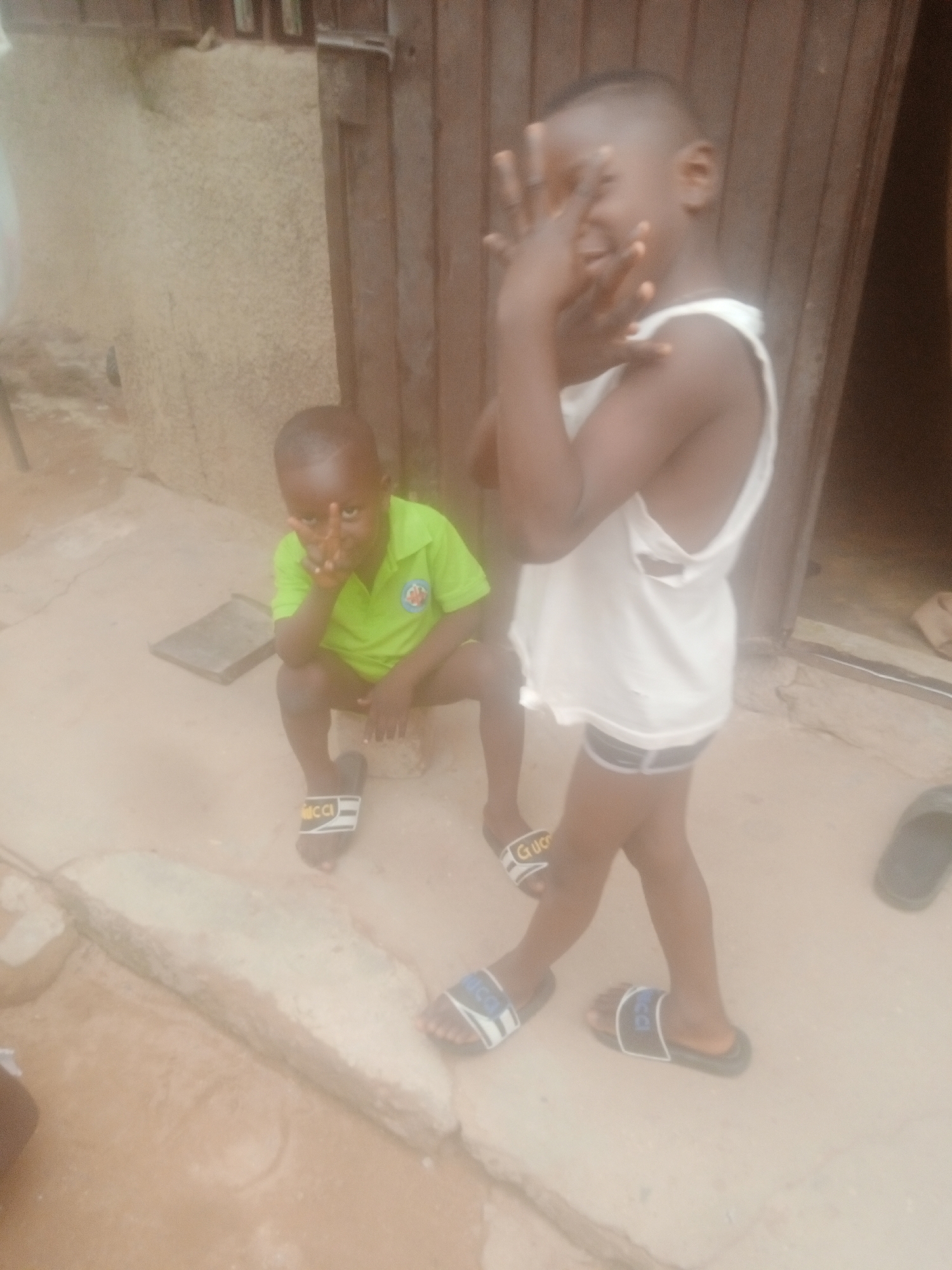 Two young black males are positioned outdoors. One stands, wearing a white tank top and dark shorts, holding their hands up near their face in what appears to be a gesture. The other sits, wearing a green collared shirt and dark shorts, also holding their hand up in a similar gesture. Both are wearing sandals. They are situated on a light-colored, possibly concrete surface, next to a wall and a wooden door. The city and location are unknown.