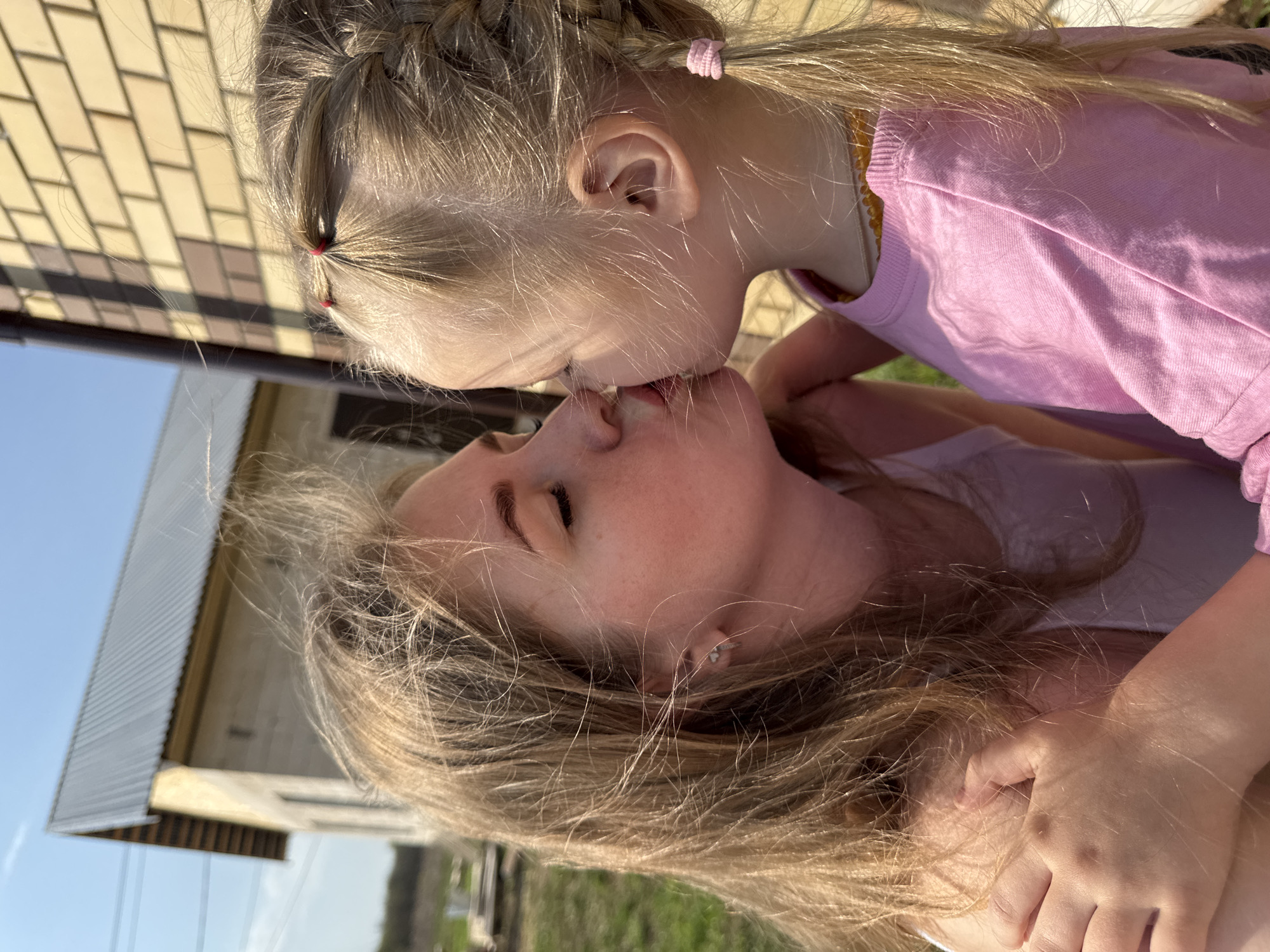 The image shows a mother and daughter kissing outdoors. The daughter, who is younger, has blonde hair styled in braids with pink hair ties. She is wearing a pink long-sleeved shirt. The mother also has blonde hair and is wearing a light-colored top. The background suggests an outdoor setting in Ферма, Russia, as the location metadata indicates. A house with a brick exterior and corrugated metal roof is partially visible. The sky is blue, suggesting a sunny day. The interaction is a loving embrace between the two subjects.
