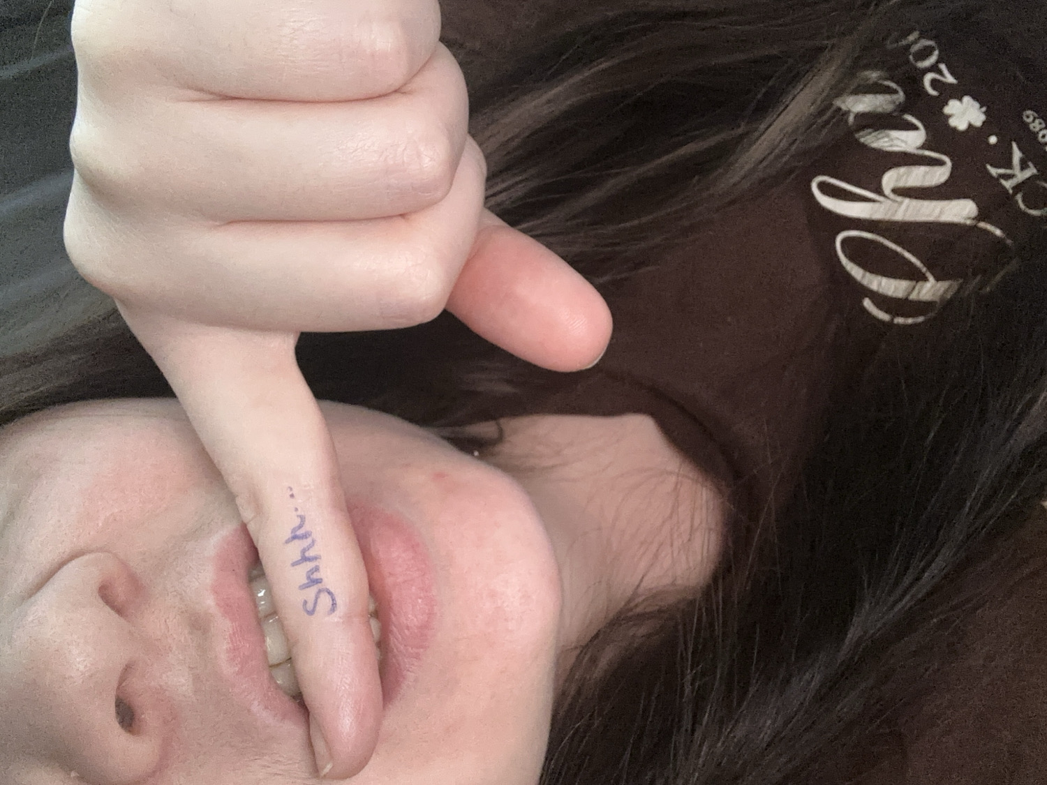 The original image, set in Chaikovsky, Russia, captures a close-up, rotated view of a person making a "shushing" gesture. A light-toned hand is prominently featured, with its index finger placed horizontally across the person's mouth. The word "Shhh..." is distinctly written in blue ink on the side of the index finger. The person's lips are slightly parted, revealing glimpses of teeth. Dark, seemingly long hair with fine strands is visible on the right side of the frame, resting against a dark brown fabric, possibly a garment. This fabric displays white graphic elements, including a stylized word that appears to end in "eye", a clear four-leaf clover symbol, and fragmented text reading "200" and "CK. * 1989". The image's rotation suggests the person might be lying down. The overall activity conveys a sense of requesting silence or sharing a secret. No specific time of day or weather is discernible. The setting appears to be indoors.