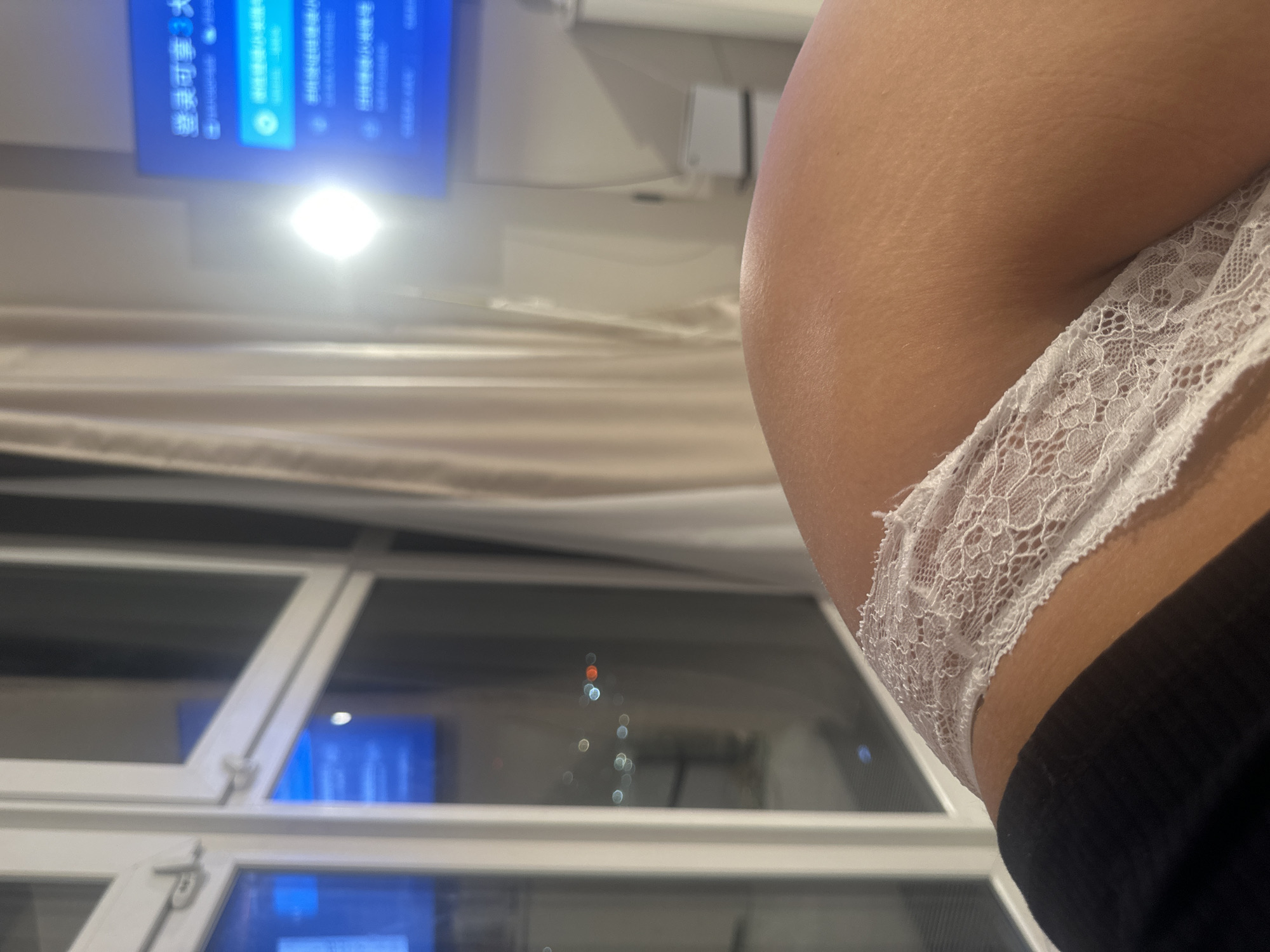 The image captures an intimate indoor scene, viewed from a low angle, focusing on a person's body and parts of the room. On the right side, a person's bare upper thigh and hip are visible, featuring white, delicate lace underwear. The skin exhibits subtle stretch marks. A dark garment, possibly clothing, is partially visible below the underwear.
The left and central parts of the image reveal the room's elements. In the upper left, a blue screen, likely a television or monitor, is displayed. Below it, a bright, intense light source creates a glare. Light-colored curtains are drawn across the mid-ground. The lower half of the image features a white-framed window, through which a dark, blurry urban landscape with colorful bokeh lights (red, orange, white) is visible, indicating nighttime.
The blue screen prominently displays Chinese characters. The most legible text is "继续可看" (Jìxù kěkàn), which translates to "Continue watching." Below this, other menu options appear, including "观看" (Guānkàn - Watch), "设置" (Shèzhì - Settings), which is highlighted, "实时" (Shíshí - Real-time), and "创建" (Chuàngjiàn - Create).
The scene suggests an evening or nighttime setting, indoors, with artificial lighting. No specific activity is discernible beyond the person being present. While the visual cues show Chinese text on the screen, the provided context indicates the location is Moscow, Russia, suggesting the viewing of Chinese media in a Russian setting.