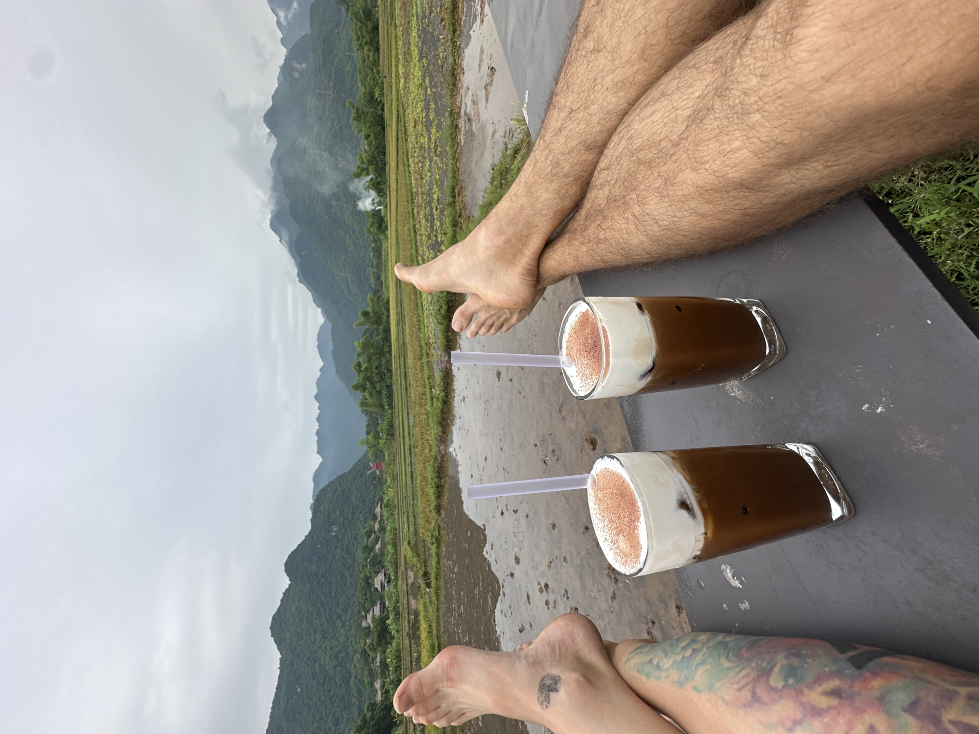 The image shows two people's legs and feet resting in front of a scenic view of terraced rice paddies and mist-covered mountains in Mai Chau, Vietnam. Two iced coffee drinks with whipped cream and cocoa powder are placed on a dark surface in the foreground. One person has a heart-shaped tattoo on their left ankle and colorful tattoos on their leg. The other person's legs are covered in hair. The background features lush green mountains, traditional stilt houses, and a cloudy sky. The overall atmosphere is one of relaxation and enjoyment of the natural beauty of Mai Chau.