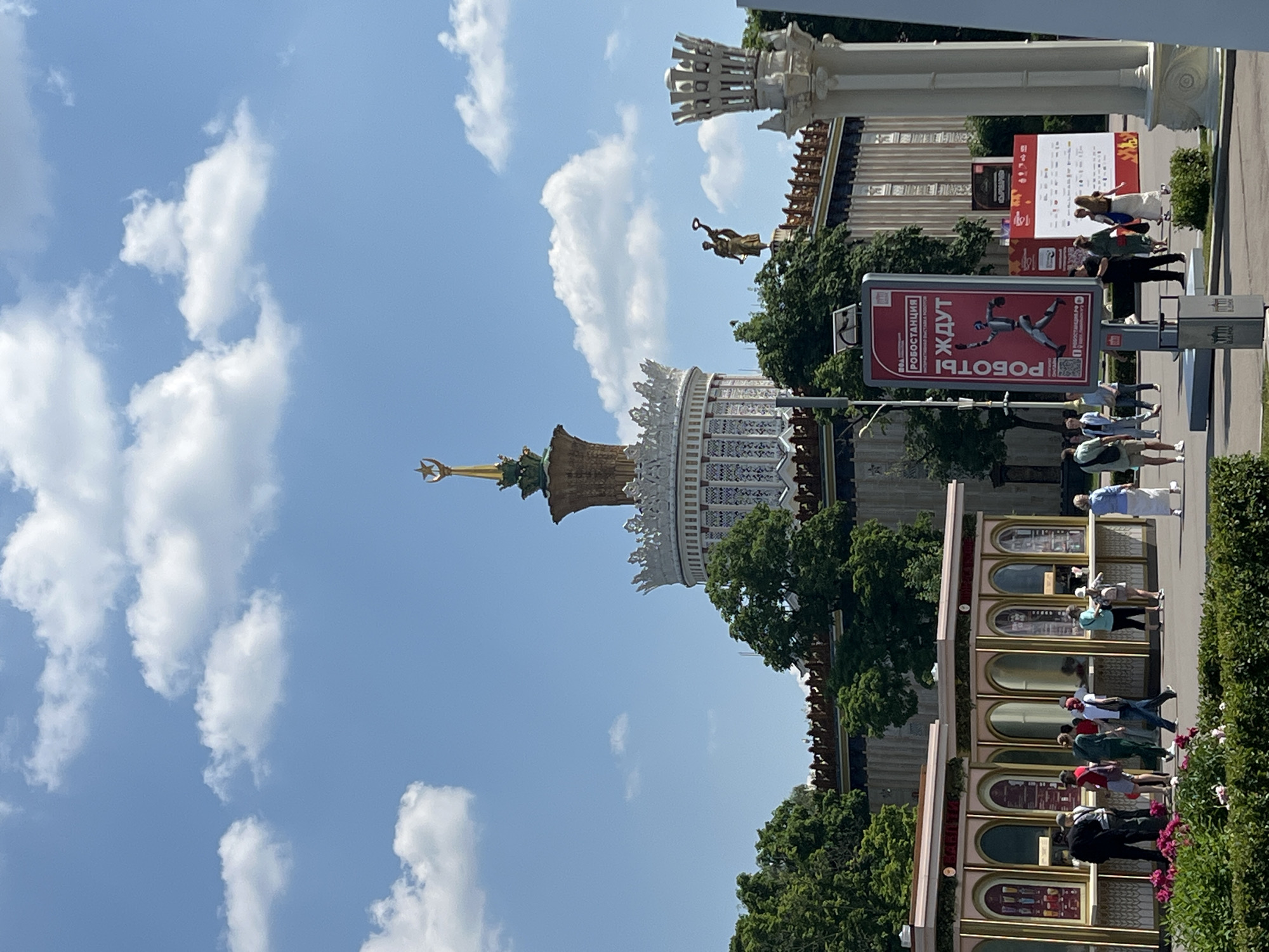This is a photograph taken on a sunny day, likely in Moscow, Russia, at the VDNKh exhibition center, as suggested by the architecture. The image showcases a grand pavilion with a distinctive gilded spire topped with a star, a symbol often associated with Soviet-era achievements. The pavilion itself is ornate, featuring a white, circular structure with intricate detailing and a crown-like cornice.
In the foreground, there are several kiosks with pink and gold trim, where people appear to be queuing or interacting with staff. A diverse group of individuals, dressed in casual summer attire, are milling about in front of these kiosks and on a paved walkway. Some are walking, while others are standing and conversing. A lush green hedge with pink flowers runs along the bottom of the frame.
To the right of the kiosks, a tall white column with decorative elements stands prominently. Further back, a large red advertising board displays an image of a robot and text in Cyrillic, indicating an exhibition or event related to robotics. More people are visible in the vicinity of this sign. Behind the foreground elements, the pavilion building stretches across the middle ground, adorned with decorative railings and surrounded by mature green trees. The sky above is a vibrant blue, dotted with fluffy white clouds, creating a picturesque backdrop. A golden statue of a woman holding a wreath is visible atop the pavilion's roofline.