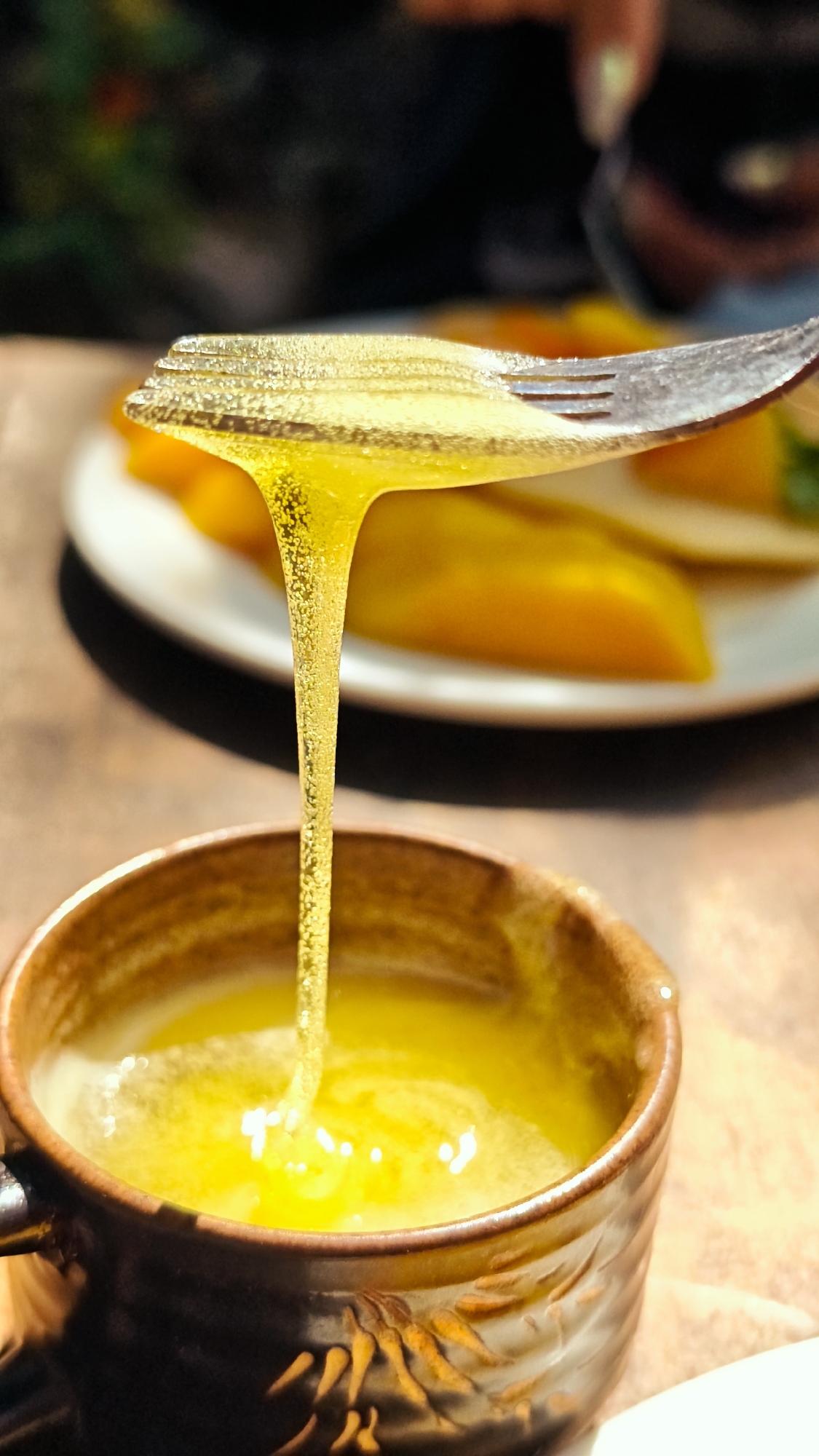 A photograph shows a close-up of honey or syrup being poured from a fork into a brown ceramic mug. The liquid is a transparent golden yellow, dripping in a thin, unbroken stream into the mug filled with a similar liquid. The fork is silver and rests on top of the mug. In the background is a plate with sliced yellow fruit. The setting appears to be an indoor dining area. Without further information, it cannot be confirmed if the image was taken in Lũng Cú Commune, Vietnam.