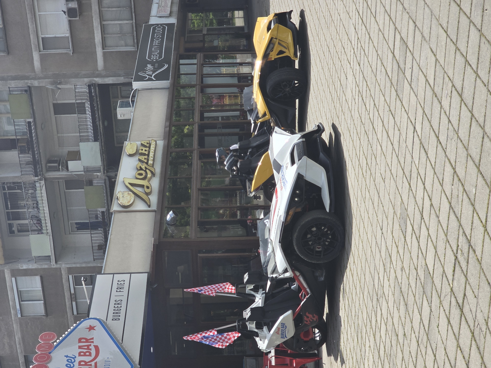 The image captures a scene in an urban environment, likely in the city of Unknown, Unknown. A row of motorized three-wheeled vehicles, possibly rental units, is parked along a sidewalk paved with patterned blocks. The vehicles are predominantly yellow and white, with black accents.
Behind the vehicles is a commercial building with a restaurant or cafe called "OZAHA FOODS" and a burger bar. The building has a large glass facade, reflecting the exterior environment. Above the storefronts, there are apartments with windows and balconies, suggesting mixed-use development.
Signage for "BEAUTY PRO STUDIO" is visible, indicating a variety of businesses in the area. The sky is visible in the background, suggesting it is daytime.
