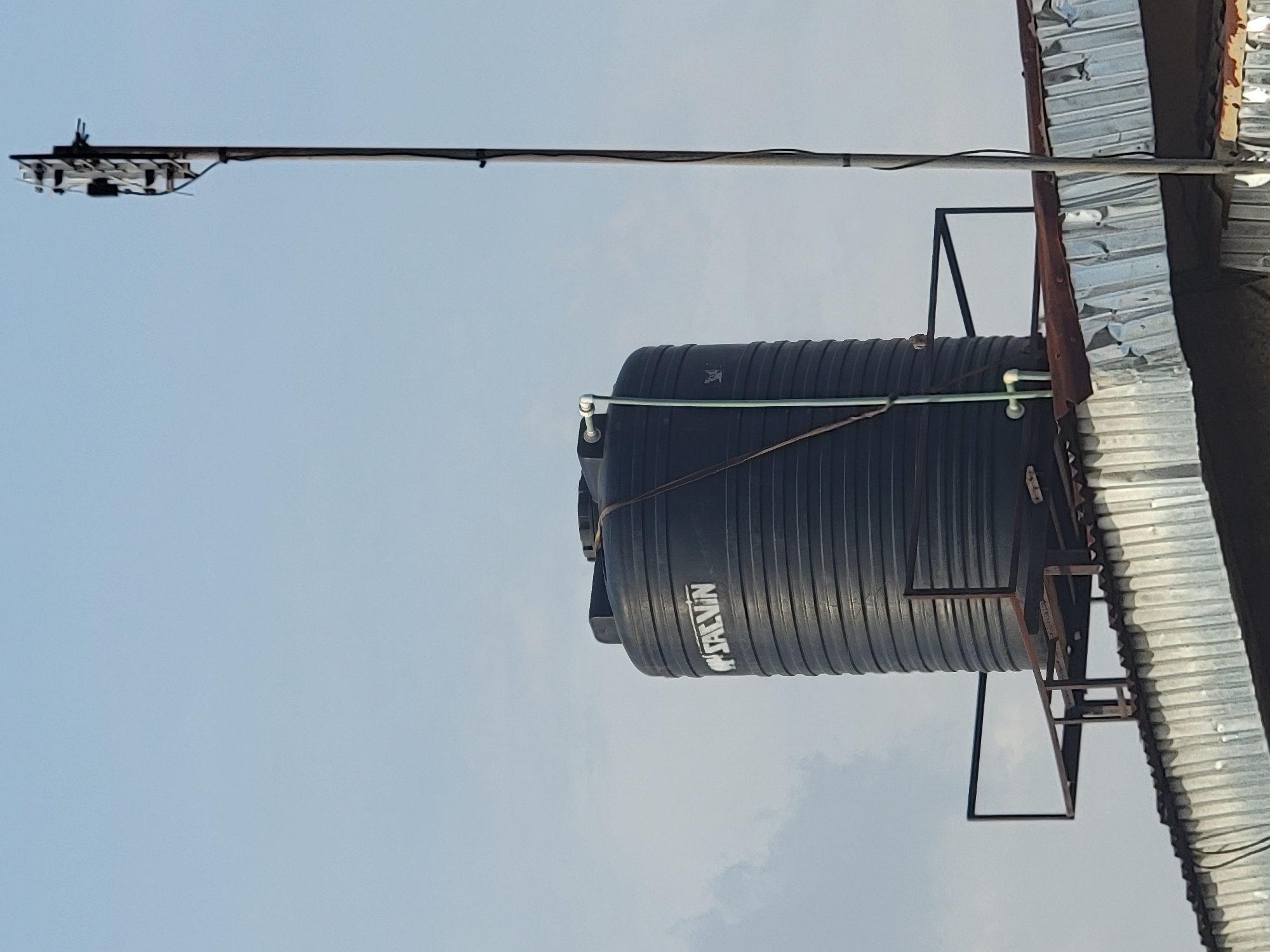 A black, cylindrical "SALVIN" water tank is mounted on a rusty, metal frame attached to a corrugated metal roof. Above the tank is a long, slender metal pole extending to the left, supporting a series of black electronic devices at its far end, possibly antennas or telecommunications equipment. The sky is overcast and light blue. The location is likely Sabon Tasha, Nigeria, as per the instructions.
