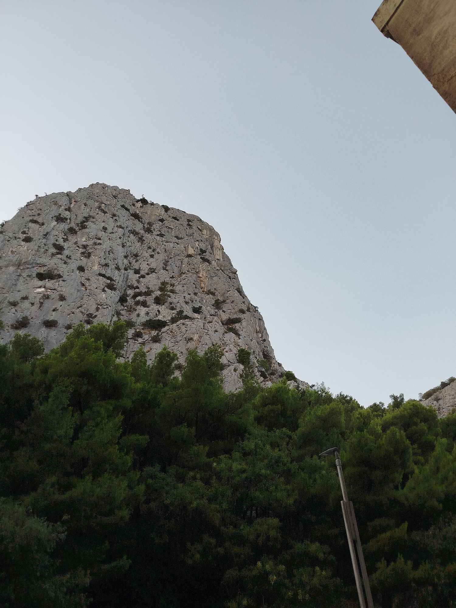 An image shows a tall, craggy mountain face in Omiš, Croatia, with a dense forest of dark green pine trees in the foreground. The sky is a pale, overcast grey. A rustic, wooden lamppost with a simple lamp fixture is positioned to the right of the frame, silhouetted against the trees and mountain. Part of a tan, possibly concrete, structure is visible in the upper right corner of the image.