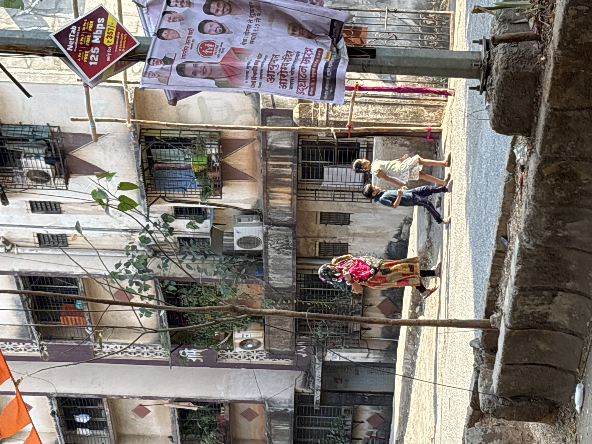 The image depicts a street scene in Ambernath, India. An older woman wearing a colorful saree and head covering walks along the sidewalk. Behind her, a boy in a dark shirt and pants and a girl in a light dress walk in the same direction. The street is lined with old, multi-story buildings with air conditioning units and barred windows. Banners advertising NetTab internet services at 125 Mbps for 357 rupees and a political event related to health care by the Bharatiya Janata Party are attached to a light pole. A partially visible orange flag is at the left. The sidewalk is cracked and in disrepair.