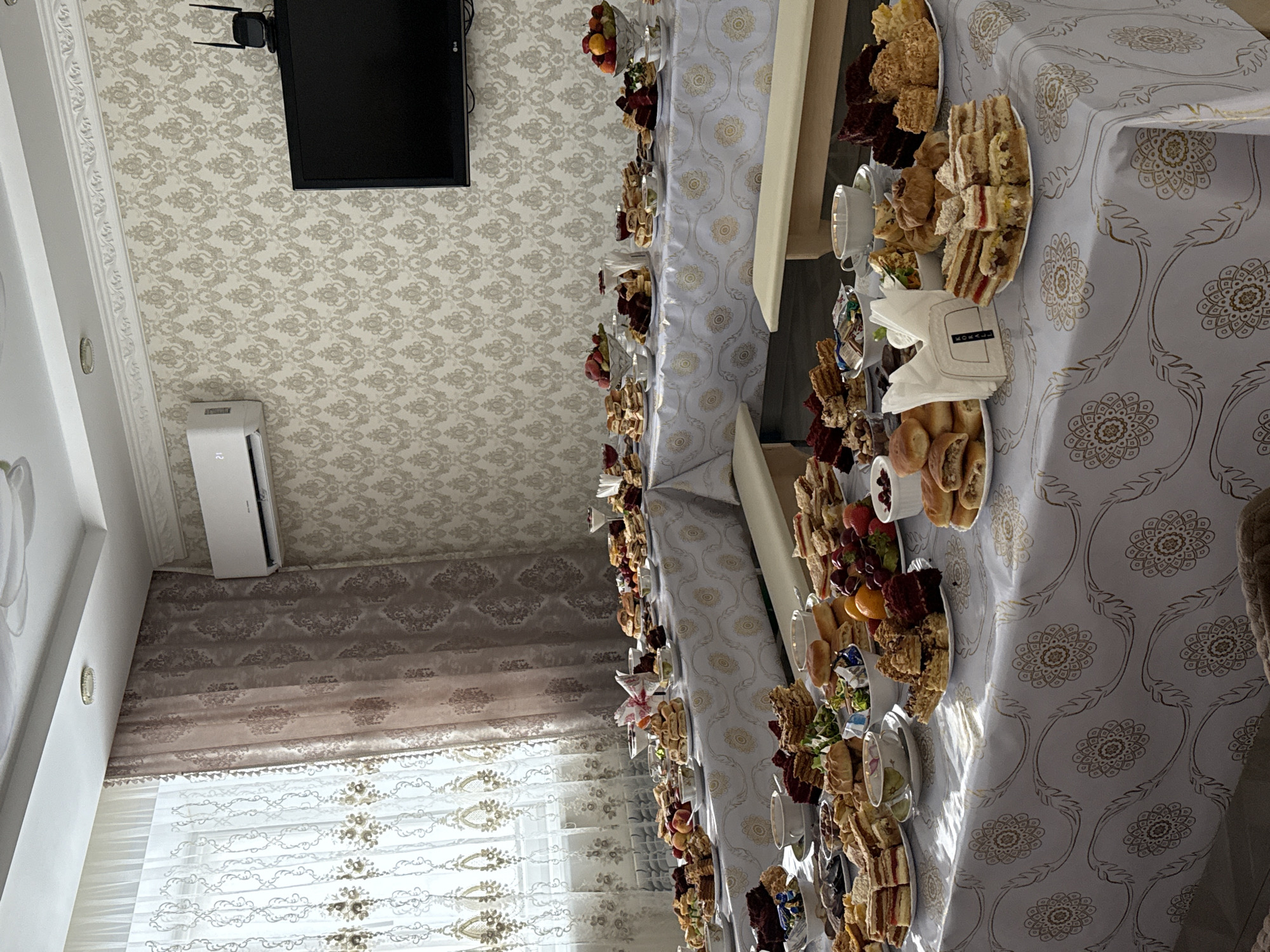 A long table laden with plates of assorted food, including sandwiches, cakes, and fruit, dominates the scene. The table is covered with a white tablecloth featuring a gold floral pattern. Napkins are stacked near the center of the table. A long, off-white cushioned bench runs along the side of the table. A window with sheer curtains and heavier patterned drapes is visible in the background. The ceiling has patterned wallpaper. A TV and an air conditioning unit are mounted on the wall. The location is Урмаево, Russia. No people are visible.