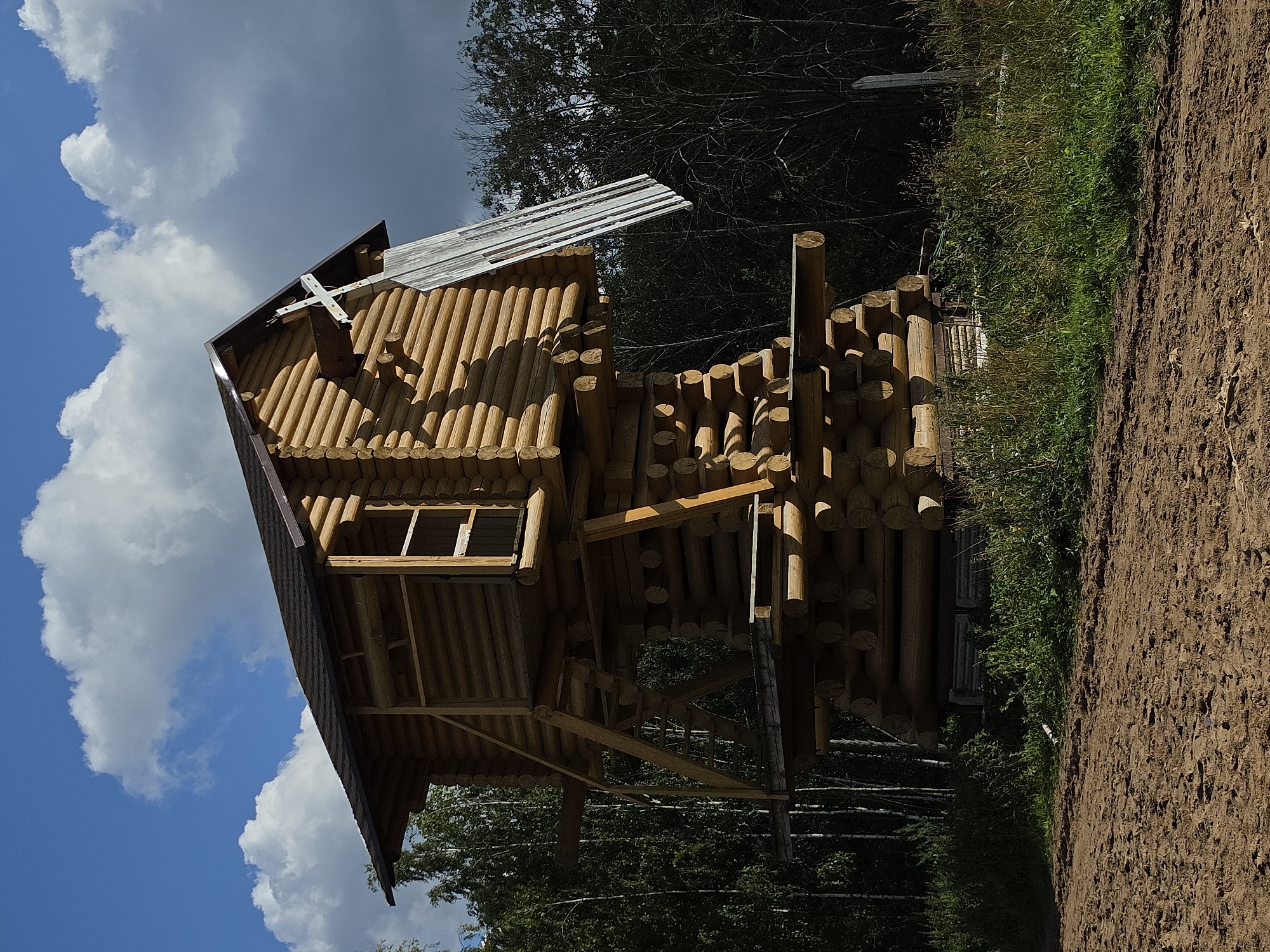 A wooden structure, resembling a stylized, multi-tiered house on stilts, stands outdoors in a rural or park-like setting. The structure is constructed from rounded logs, stacked horizontally. The lowest tier is the widest and appears to be a foundation, with logs supporting successive, narrower levels. A simple wooden ladder extends from the ground to a platform on the second level. The uppermost level is a small, enclosed cabin with a pitched roof. A white, cross-shaped element is attached to the roof, angled upwards. The building is situated amidst dense, dark green trees, with some lighter green foliage and tall grasses in the foreground. The ground is a reddish-brown dirt path with visible tire tracks or footprints. The sky above is a vibrant blue, punctuated by large, fluffy white clouds and some darker, shadowed clouds. The overall impression is one of rustic architecture in a natural environment under a dynamic sky. The specific location within Peterhof, Russia, cannot be determined from the image alone, as the image lacks any identifiable landmarks or signage characteristic of that city.