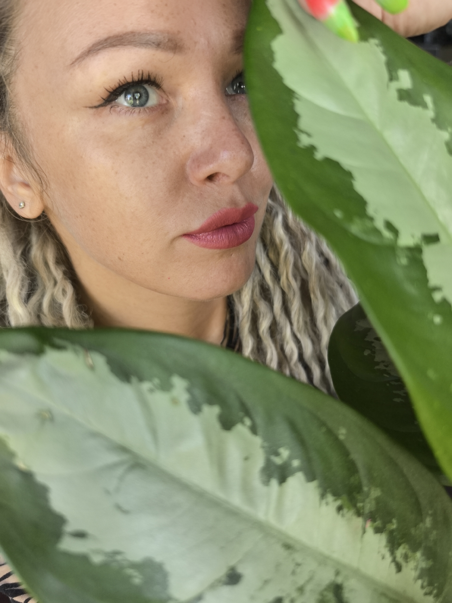 The image shows a woman's face partially obscured by large, variegated green leaves. Her skin is fair with freckles, and she has light-colored, braided hair. She has light blue eyes with dark eyeliner and pink lipstick. A small earring is visible in her ear. The leaves are in the foreground, with the woman's face appearing behind them. The background is not visible. There is no evidence in the picture suggesting a location in Noviy, Russia.