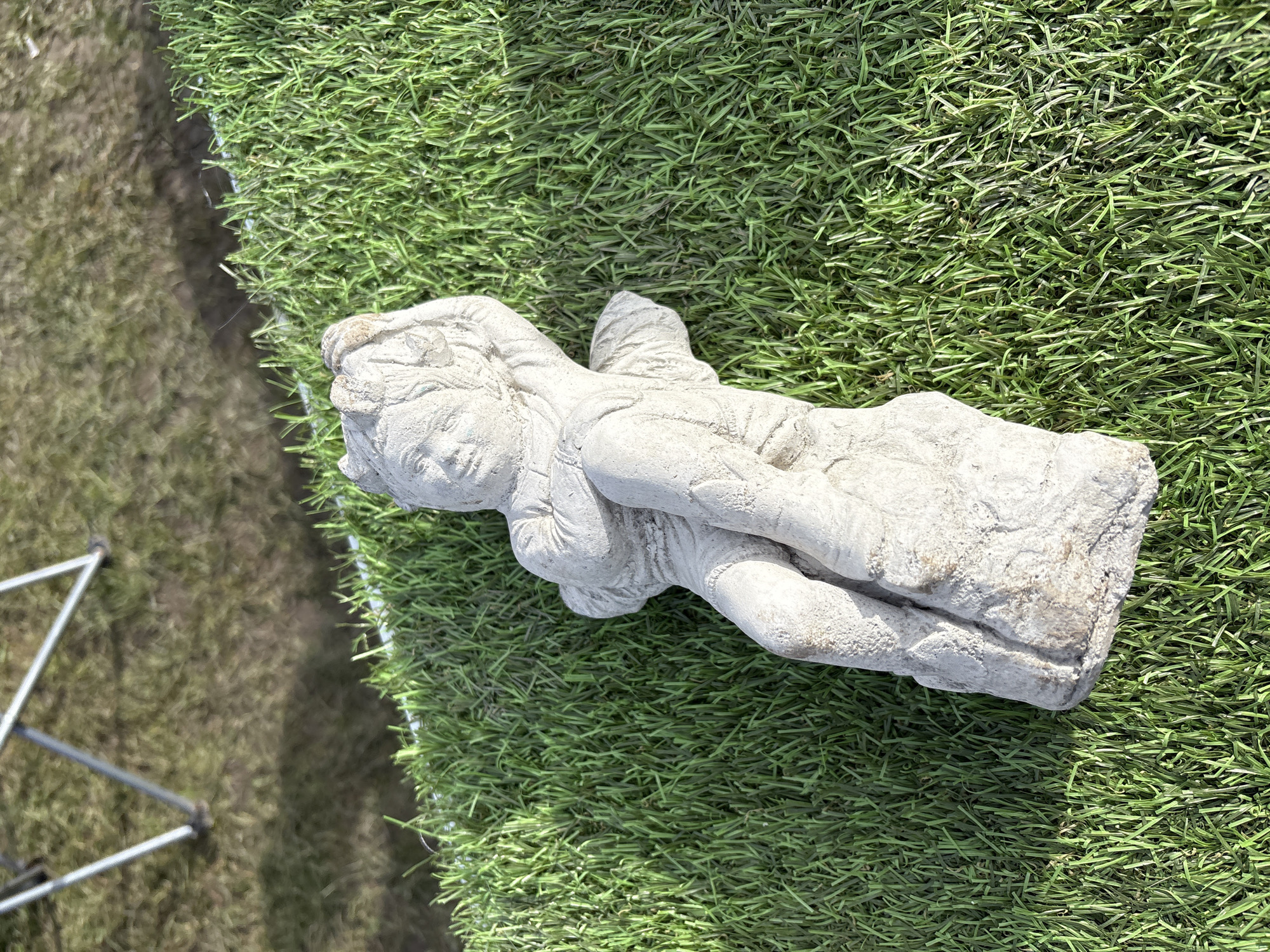 The image shows a stone statue of a person laying on its side, set on artificial grass. The statue is a light gray color, with visible weathering and texture. A tripod is visible in the lower left corner. The scene is located outdoors. The location in the city of Chirk, United Kingdom, is not discernible from the image.