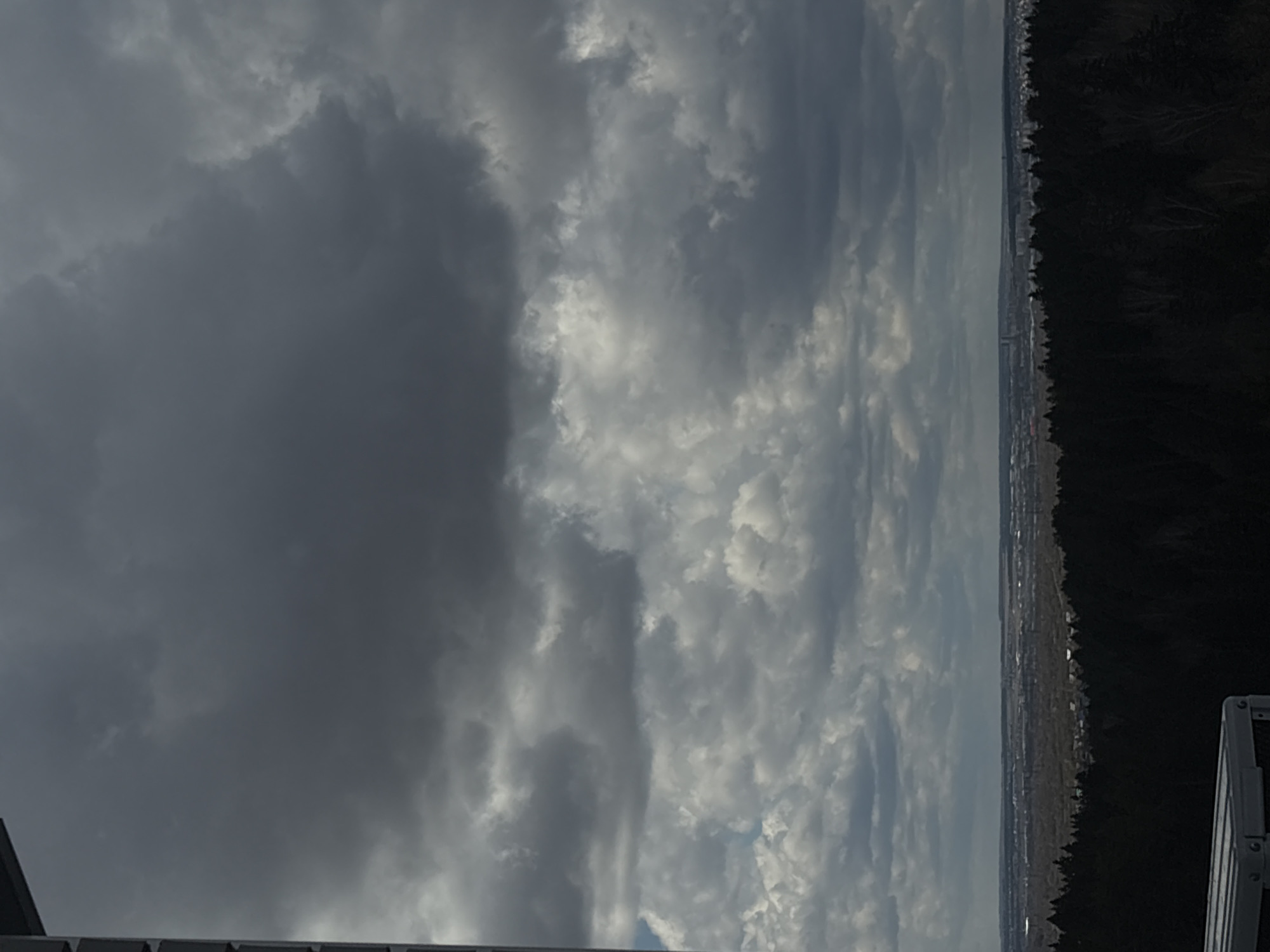 The image presents a wide, panoramic view dominated by a dramatic sky over a distant landscape, likely captured from an elevated position. The sky is filled with a mixture of clouds, varying in density and color. On the left side, large, heavy masses of dark grey clouds loom, suggesting a somber or stormy atmosphere. Moving towards the right, the clouds become lighter, more broken, and patchy, with areas of bright white indicating sunlight breaking through or reflecting off them.
Below the expansive sky, a distant horizon stretches horizontally across the lower portion of the frame. On the far right, a dark, dense treeline or forest forms a distinct boundary, contrasting with the lighter sky and landscape. Between the forest and the cloud base, a faint, hazy, and sprawling urban or suburban area can be discerned, consistent with a city or town like Izhevsk, Russia, in the distance.
In the bottom-left corner, a dark, angled structure, possibly a part of a building's roof, is partially visible. In the bottom-right corner, a section of a metal railing or similar architectural feature is present. These elements suggest the photograph was taken from an elevated vantage point, such as a high-rise building or balcony. The overall impression is one of an overcast or potentially pre-stormy day, with the light conditions indicating daytime. There is no visible text in the image.