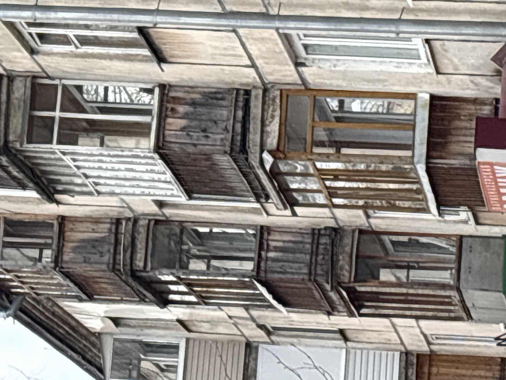The image shows a three-story building in Izhevsk, Russia. The building's facade is weathered and appears to be constructed from concrete panels. The windows on the upper two floors have small balconies with partially collapsed or missing railings. The windows are varying in style and condition, some are boarded up. The building has an aged appearance, suggesting it may be abandoned or in disrepair.